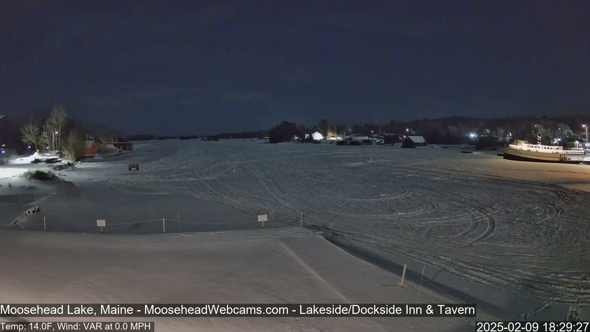 A frozen lake at night, under a dark sky, shows a few buildings and a boat on the ice, with tire tracks visible on the frozen surface.