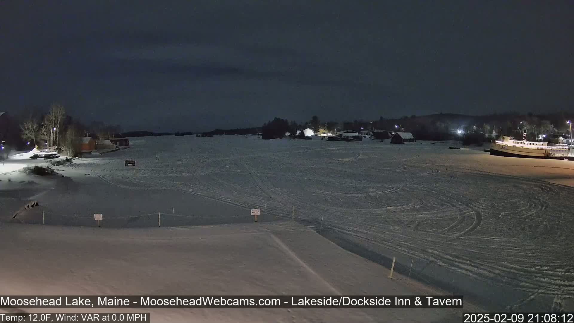 A frozen lake at night is viewed from a snowy shoreline, with buildings and a boat visible in the distance under a dark sky.