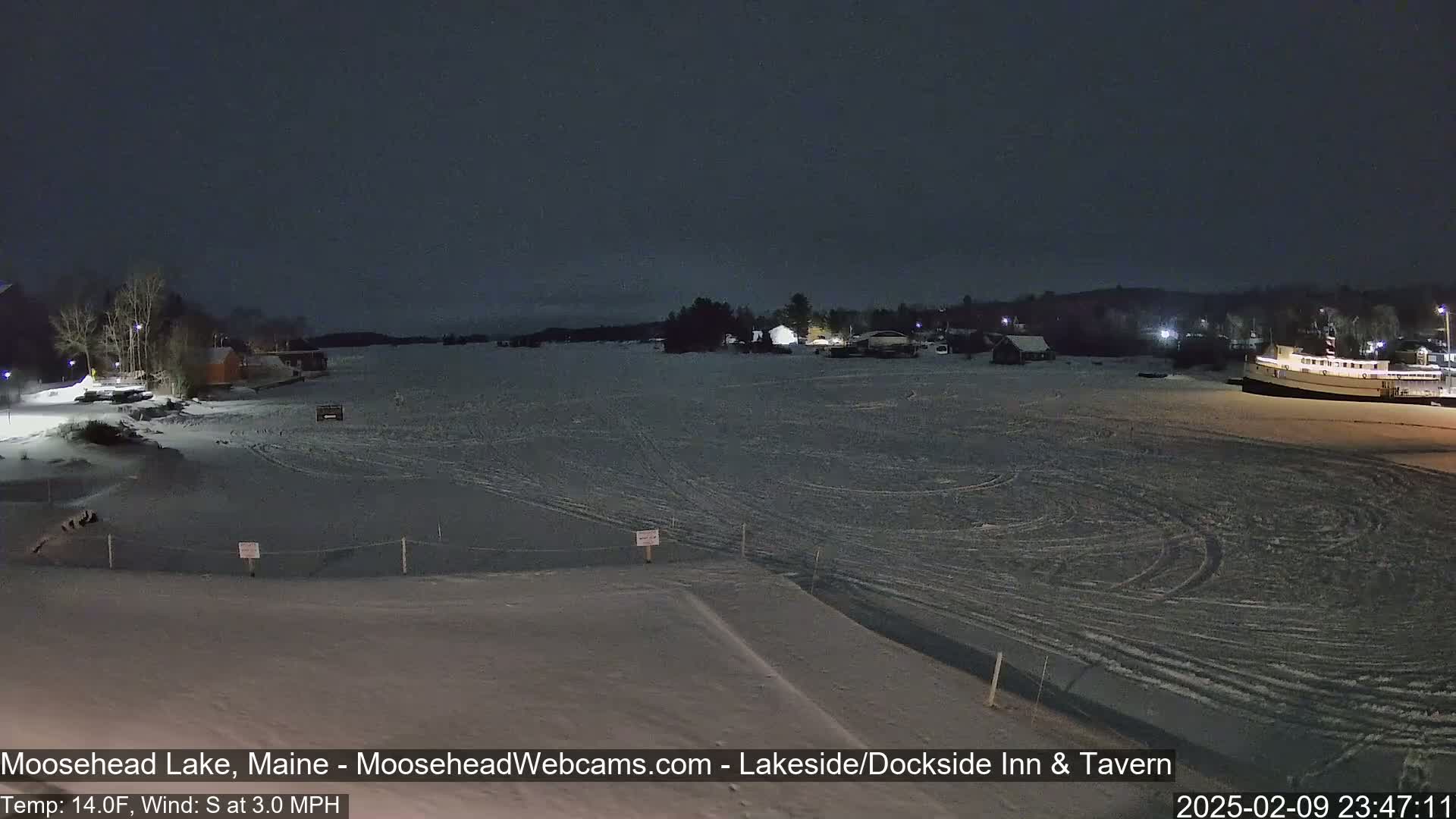 A nighttime view of a snow-covered frozen lake with buildings and a boat visible in the distance under a dark sky.