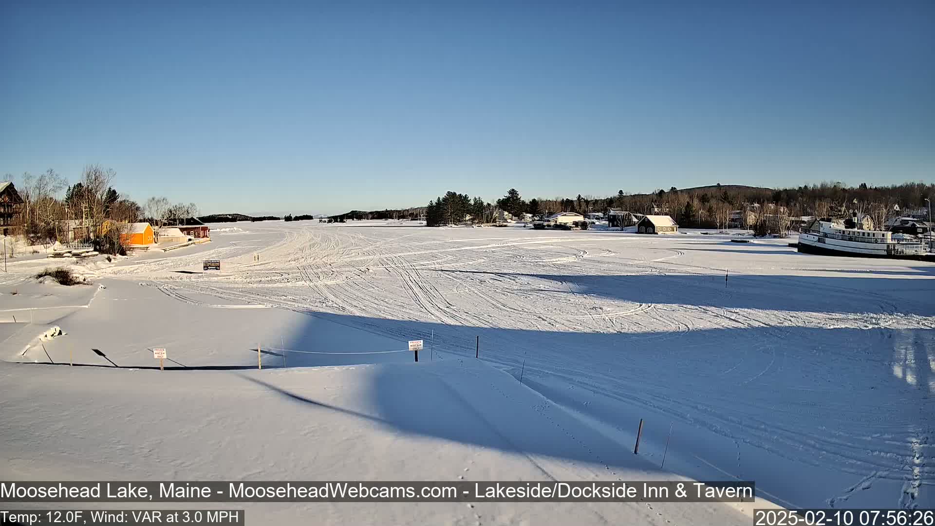 A snow-covered frozen lake, under a clear blue sky, features several buildings and a boat on its shore.