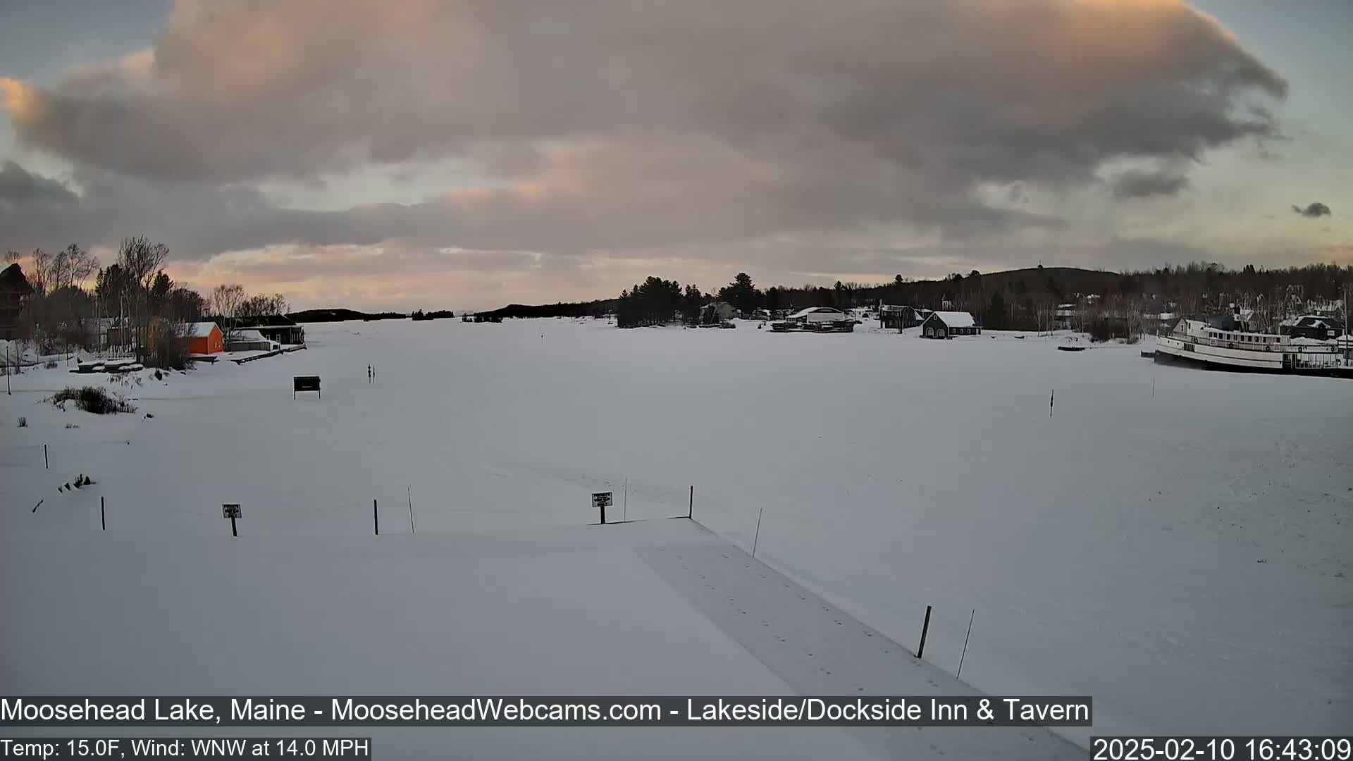 A frozen lake with snow-covered land and buildings, under a cloudy sky with a hint of pink at the horizon, in windy 15°F weather.