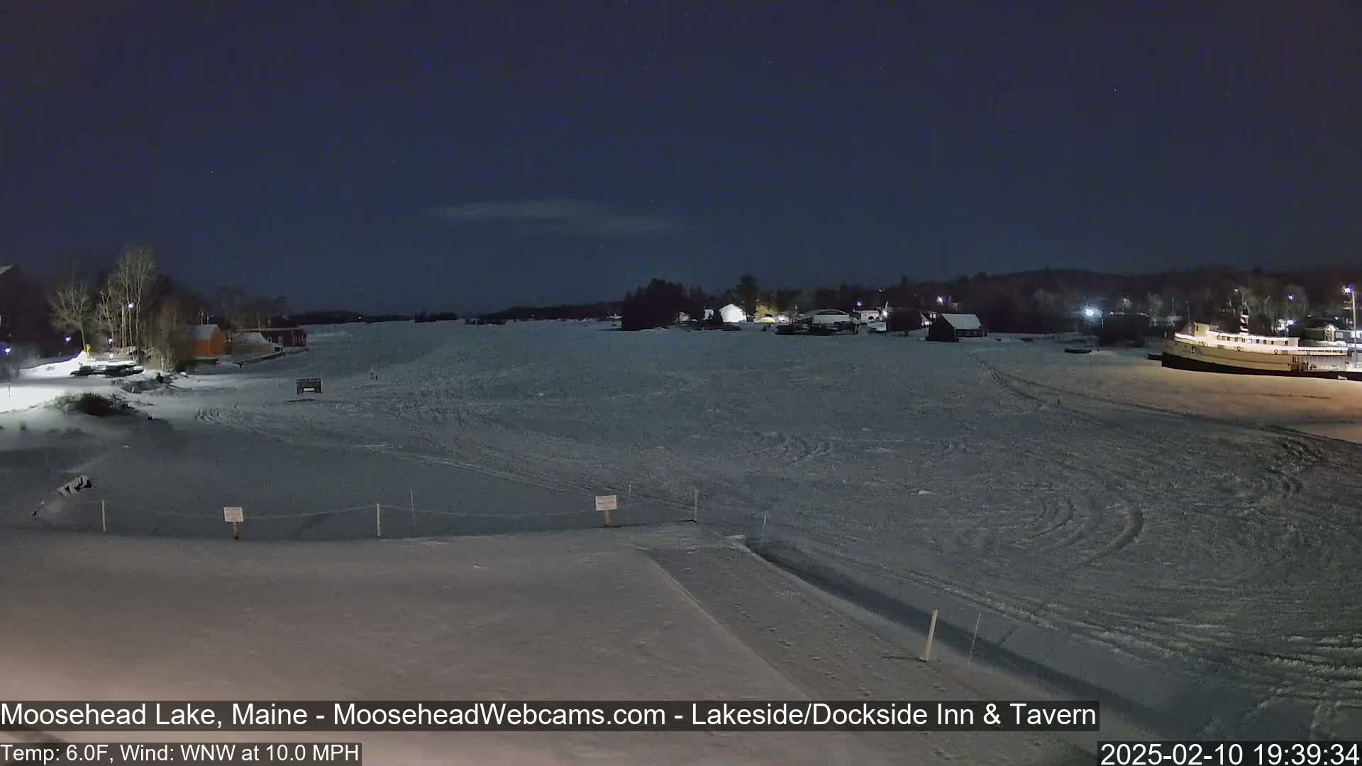 A snow-covered frozen lake at night, with a few buildings and a boat visible in the distance under a dark sky.