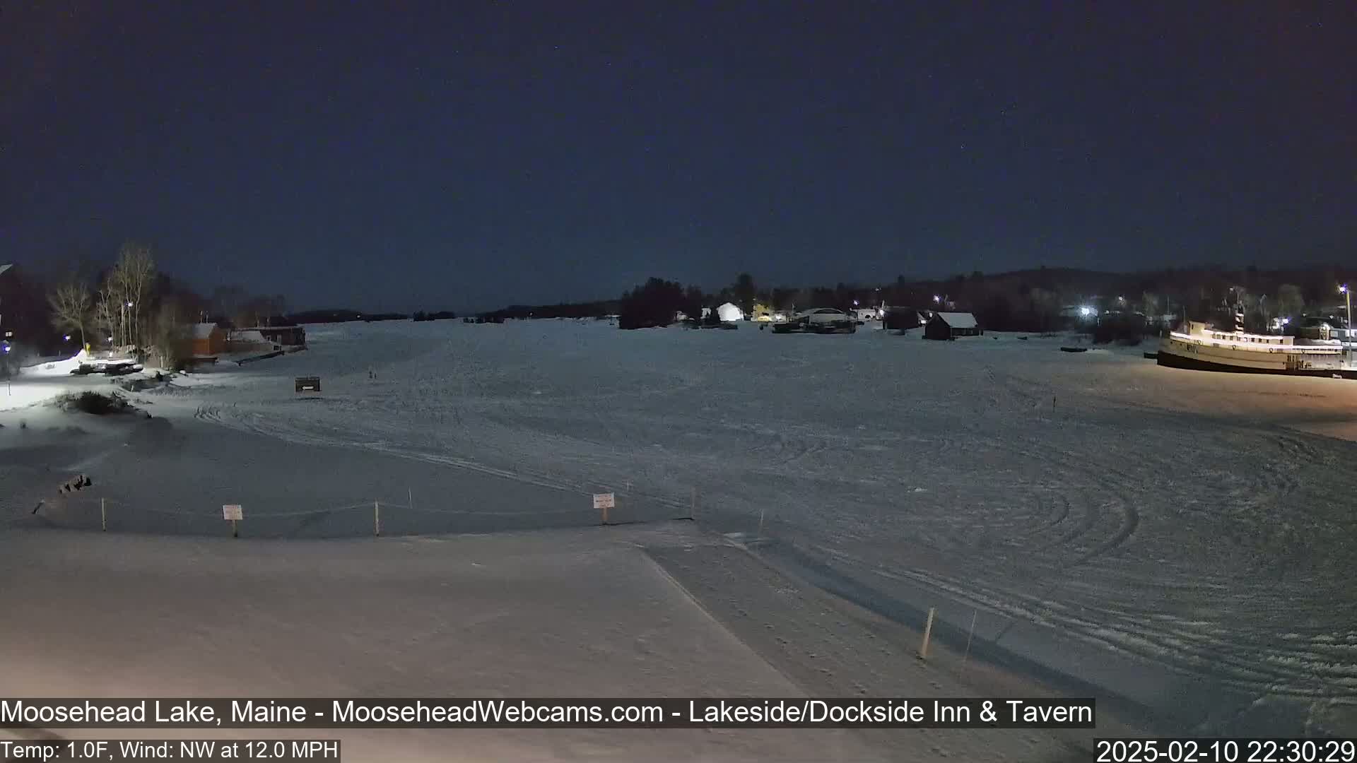 A snow-covered frozen lake at night, under a dark sky, with buildings and a boat visible in the distance, in a 1°F temperature with a 12 mph northwest wind.