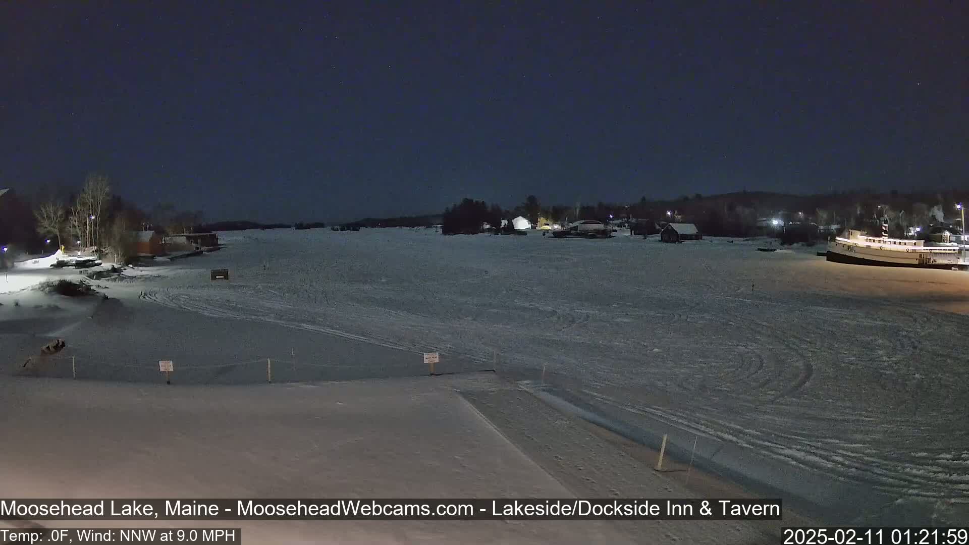A frozen lake at night, under a dark sky, shows a few buildings and a boat, with snow on the ground and a light wind.