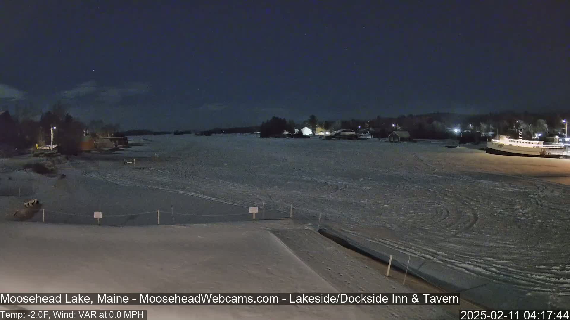 A frozen lake at night, under a dark sky, shows a few buildings and a boat docked on the ice.