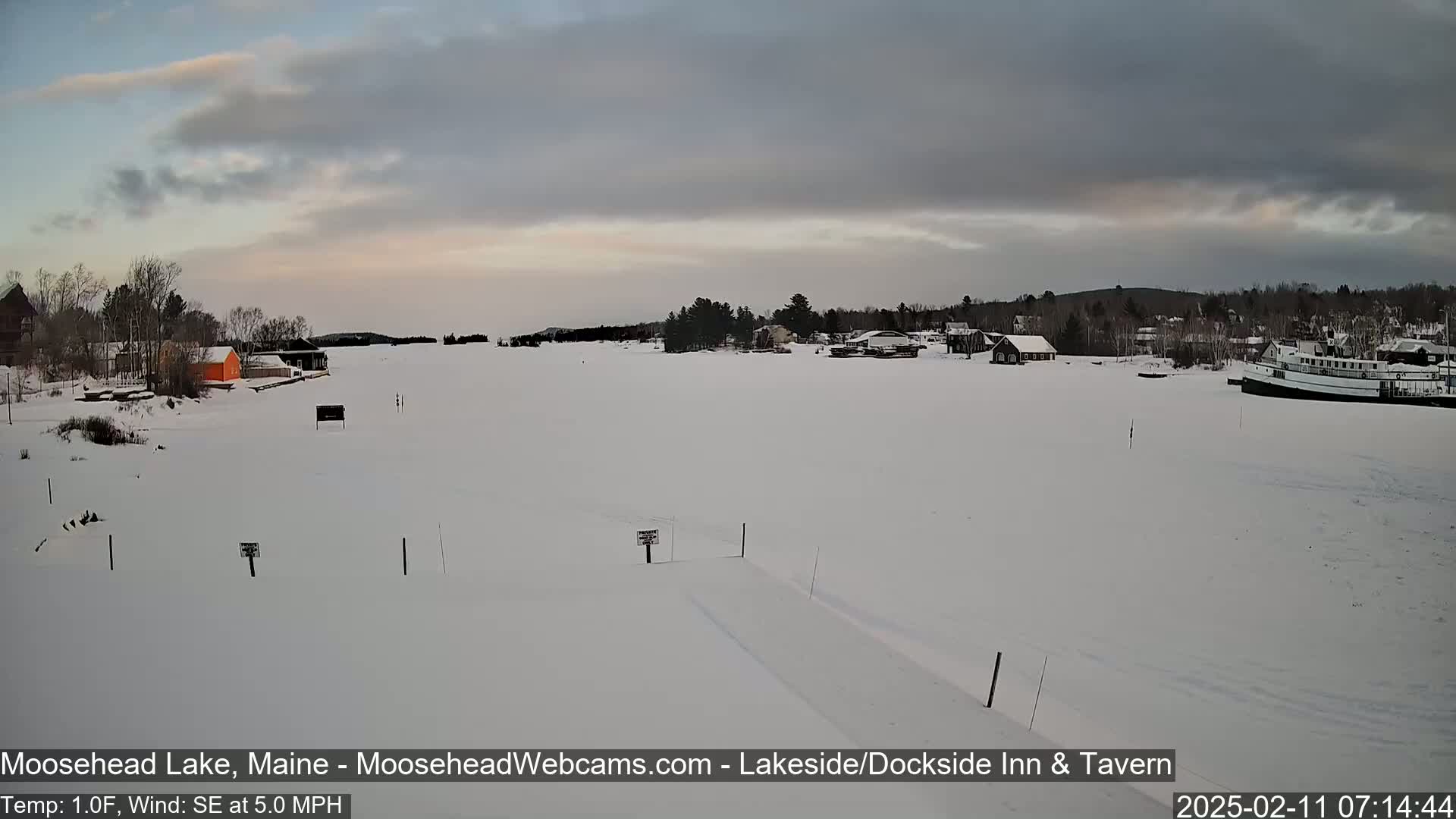 A snow-covered lake with buildings and a boat visible in the distance under an overcast sky.