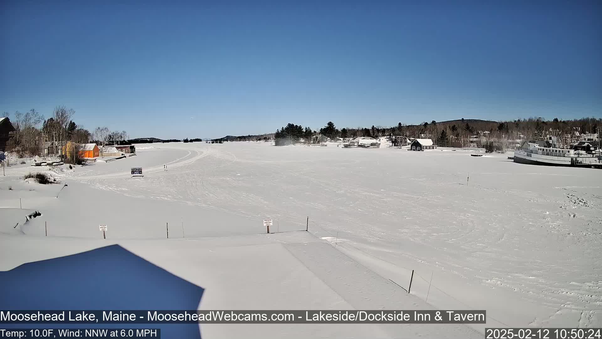 A snow-covered frozen lake stretches out under a clear blue sky, with a few buildings and a boat visible on the shore.