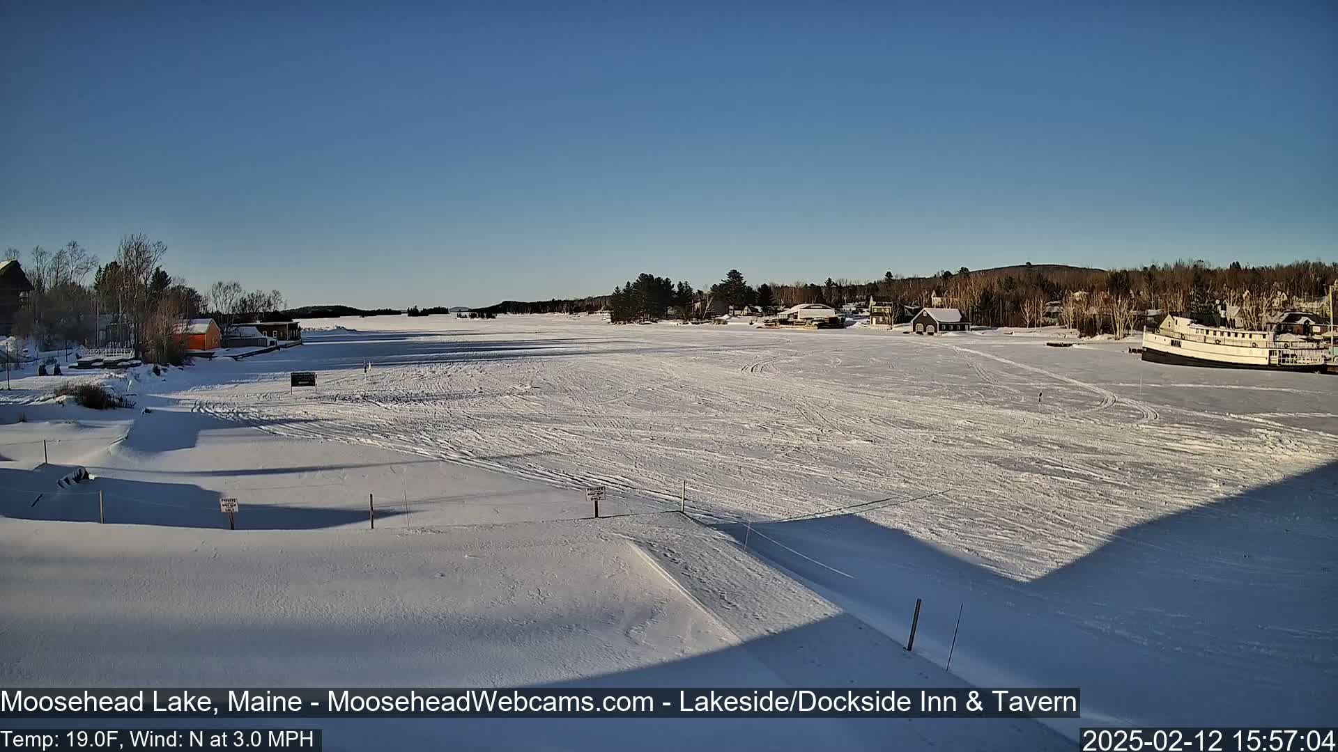 A frozen lake under a clear blue sky is seen from a snowy shoreline, with a few buildings and a boat visible in the distance.