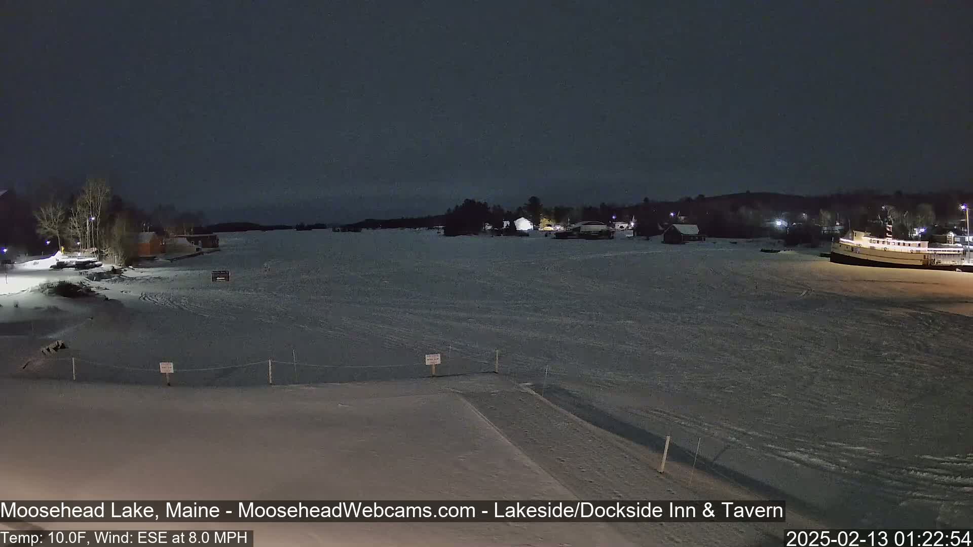 A frozen lake at night, under a dark sky, shows a few buildings and a docked boat along its shore, with snow covering the ground.