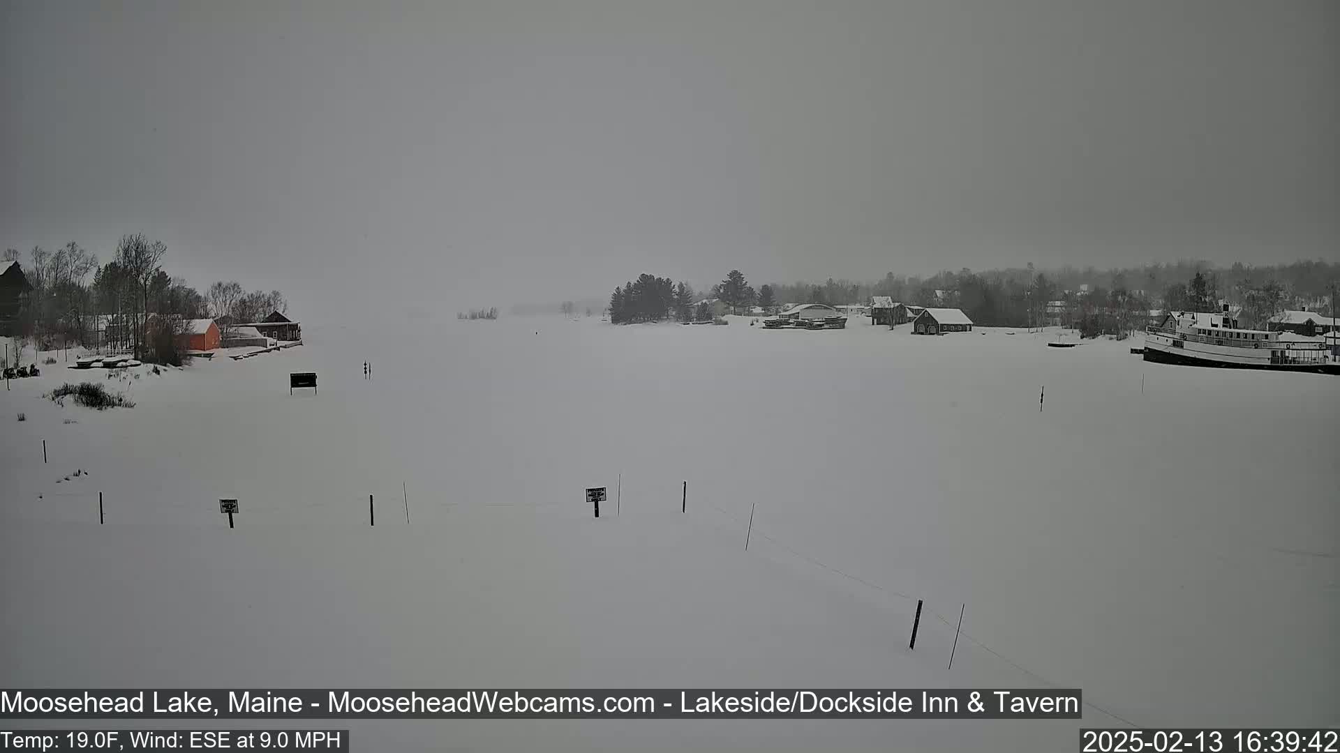A snow-covered lake scene with buildings and a boat in the distance under overcast skies.