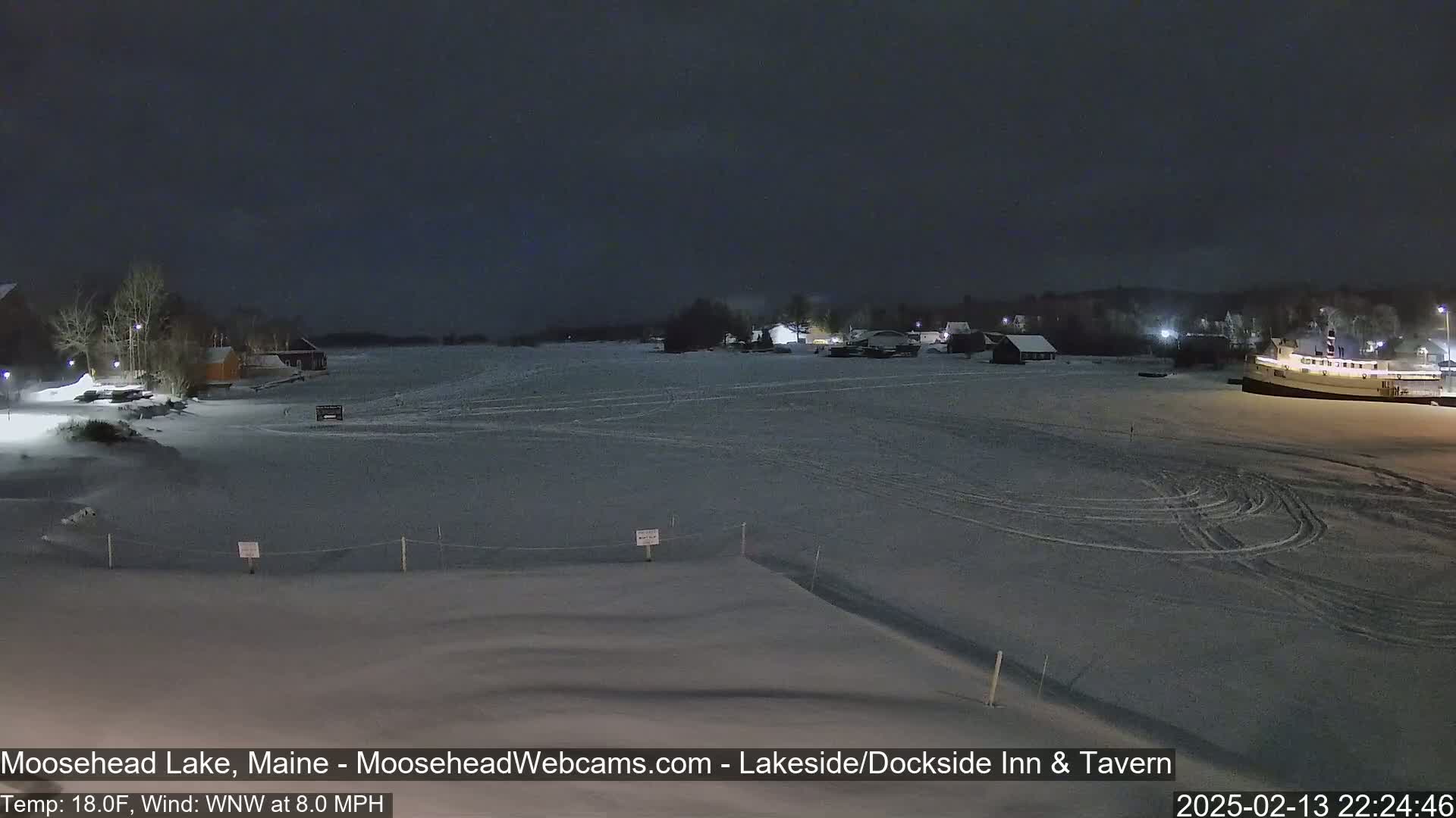 A snow-covered frozen lake at night, with buildings and a boat visible in the distance under a dark sky, with a light wind.