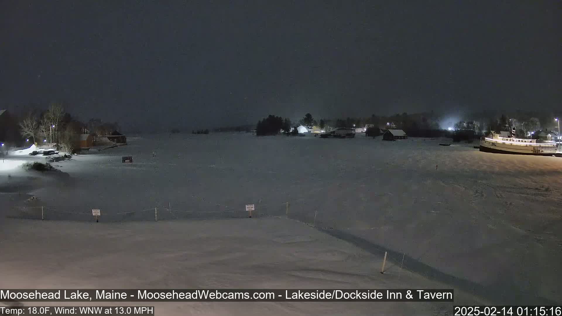 A snow-covered lake at night, with buildings and a boat visible in the distance under a dark, possibly snowy sky.