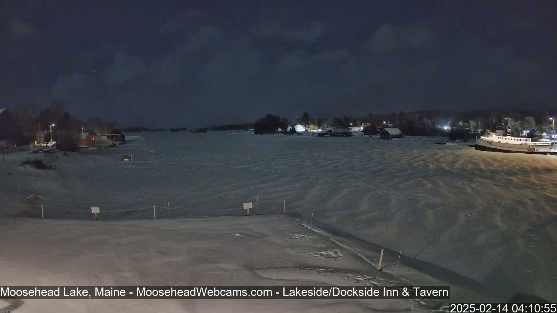 A snow-covered shoreline at night overlooks a mostly frozen lake with a boat and buildings visible in the distance under a dark, clear sky.