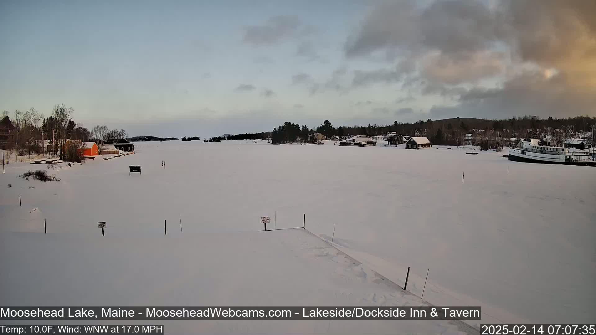 A snow-covered lake with buildings and a boat on the far shore under a cloudy sky.