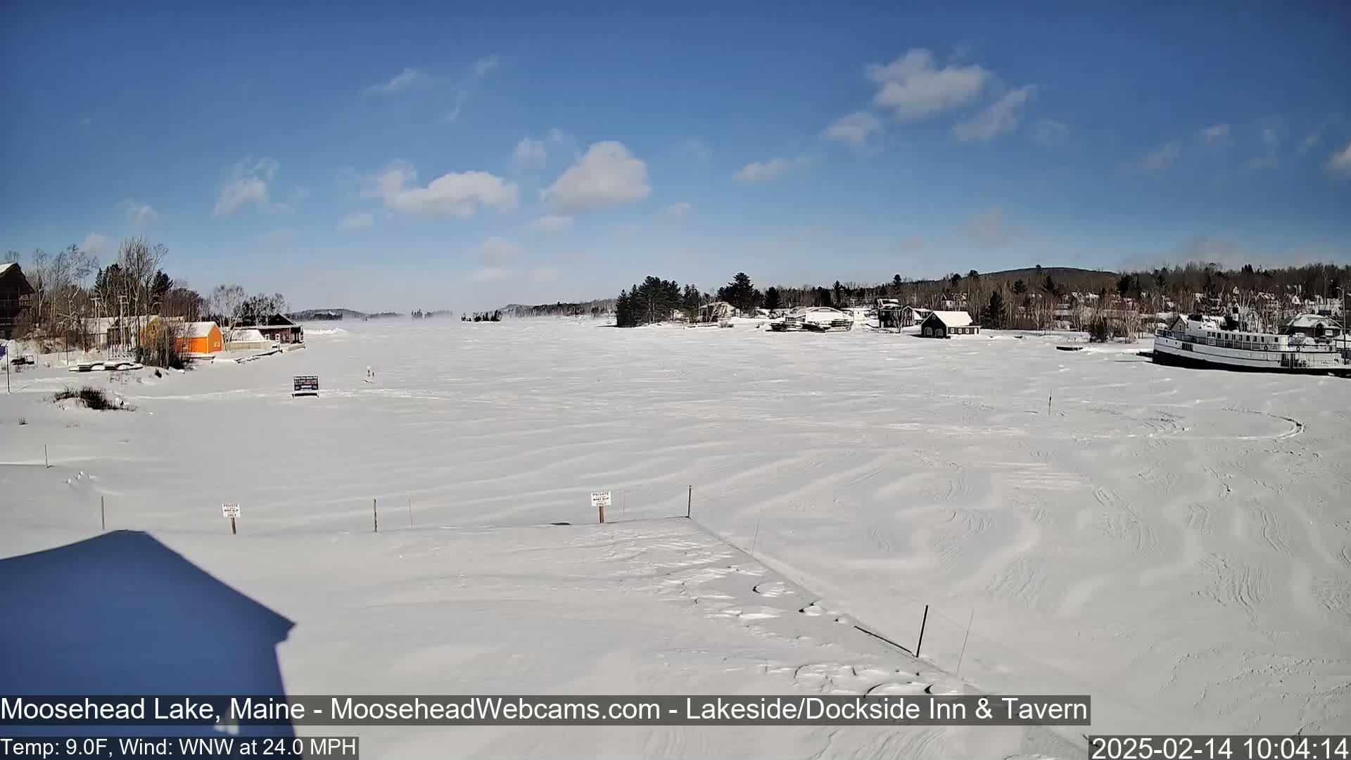 A frozen lake, covered in snow with wind-swept patterns, features buildings along the shore under a mostly clear blue sky.