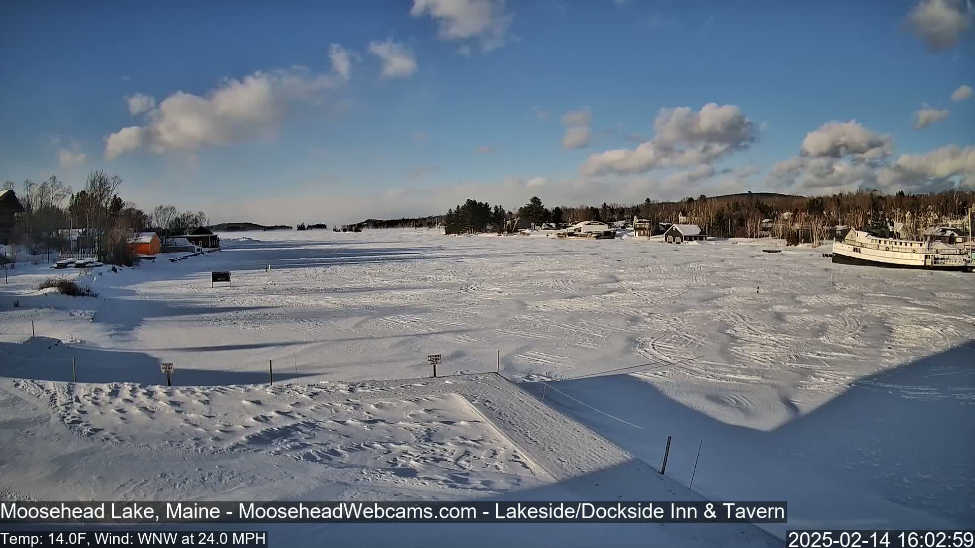 A mostly sunny, snowy scene of a frozen lake with buildings and a boat along the shore, under a partly cloudy sky with a brisk wind.