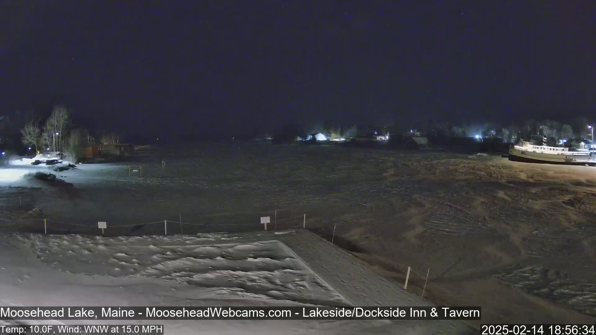 A nighttime view of a snow-covered lake shore with a few buildings and a boat visible in the distance under a dark sky, with a wind of 15 mph.