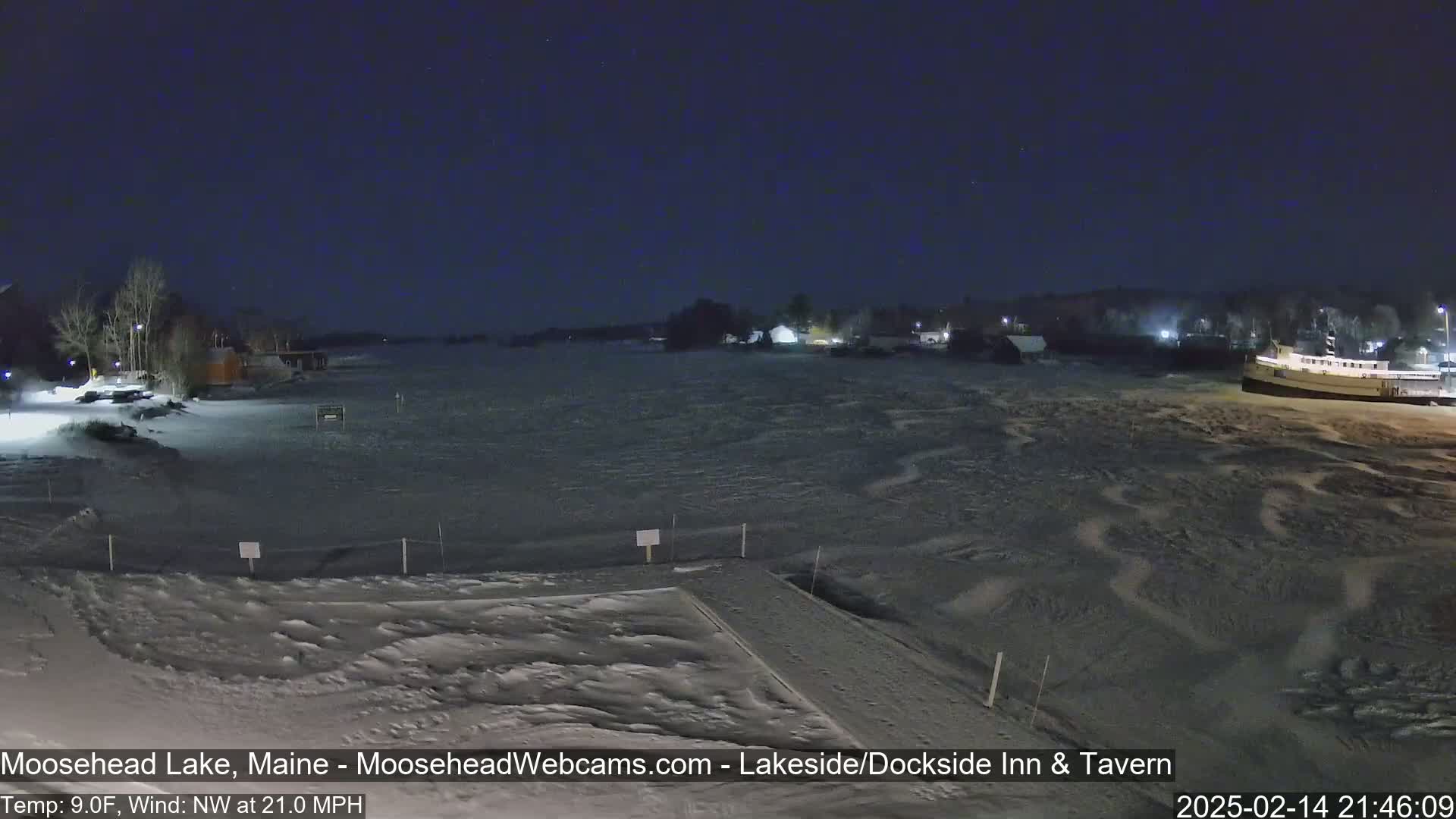 A nighttime view of a snow-covered lake and shoreline with buildings in the distance under a dark, clear sky, with a strong northwest wind.