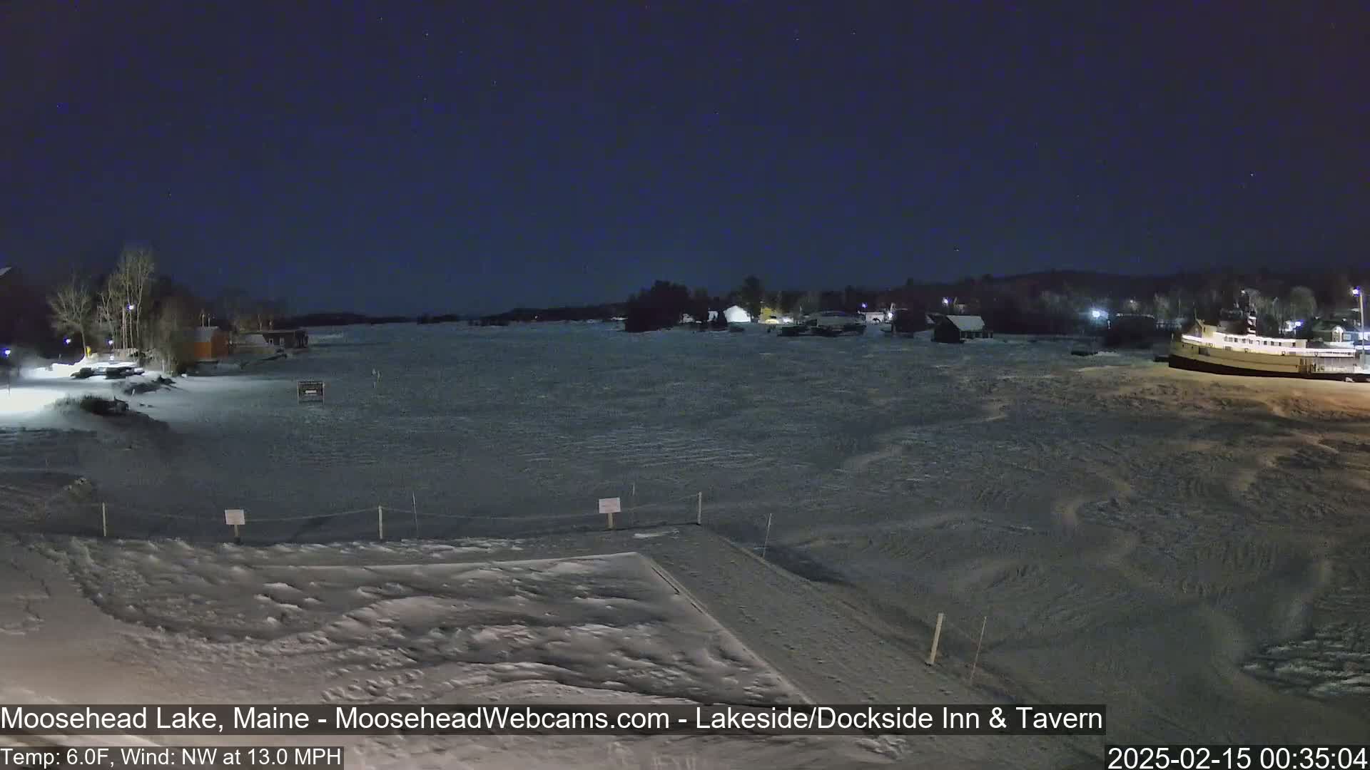 A snow-covered lake at night under a dark, starry sky, with buildings and a boat visible in the distance, a northwest wind blowing at 13 mph, and a temperature of 6 degrees Fahrenheit.