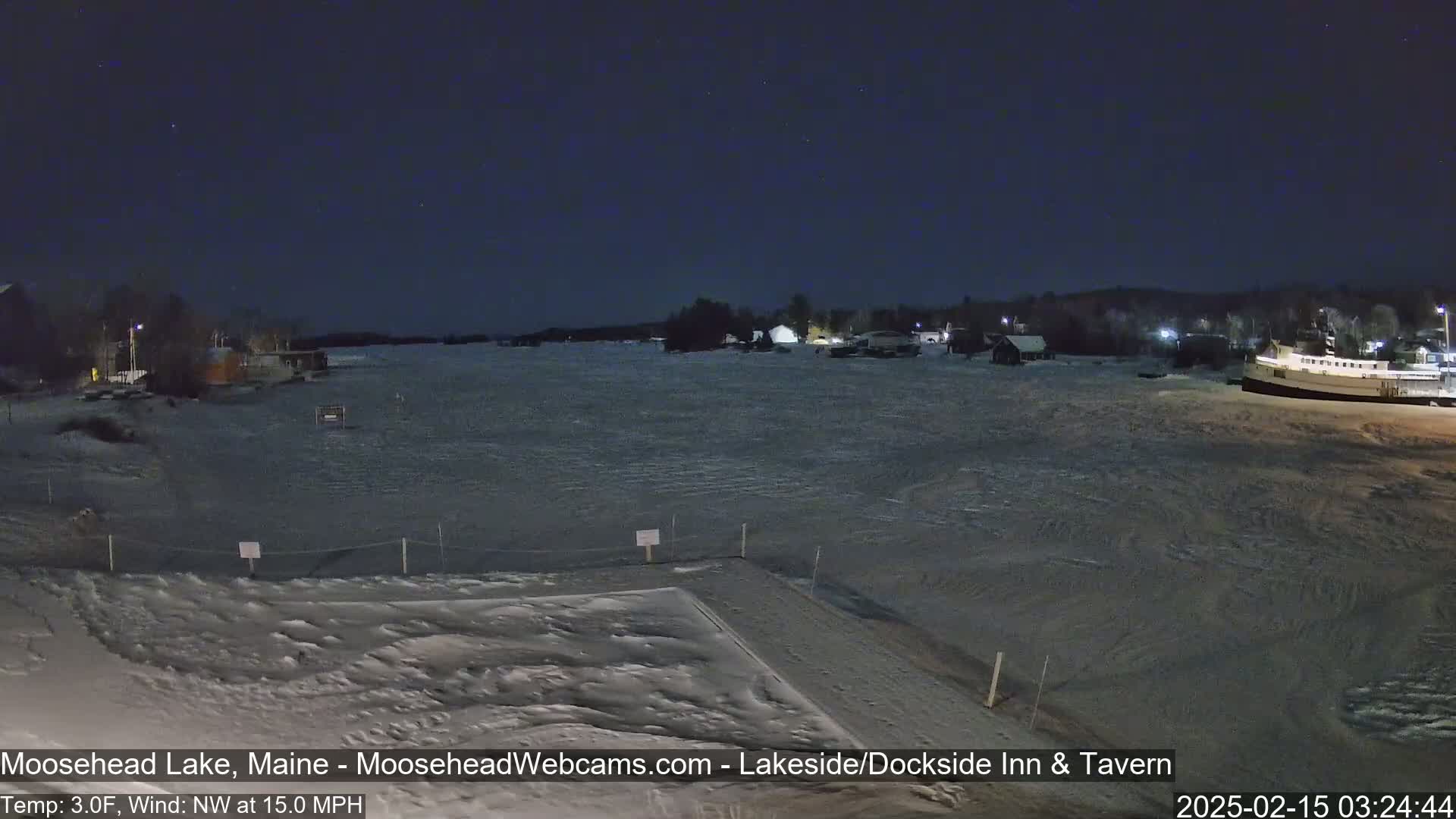A snow-covered frozen lake at night, under a dark sky with some stars visible, shows a few buildings in the distance with a northwest wind at 15 mph and a temperature of 3 degrees Fahrenheit.
