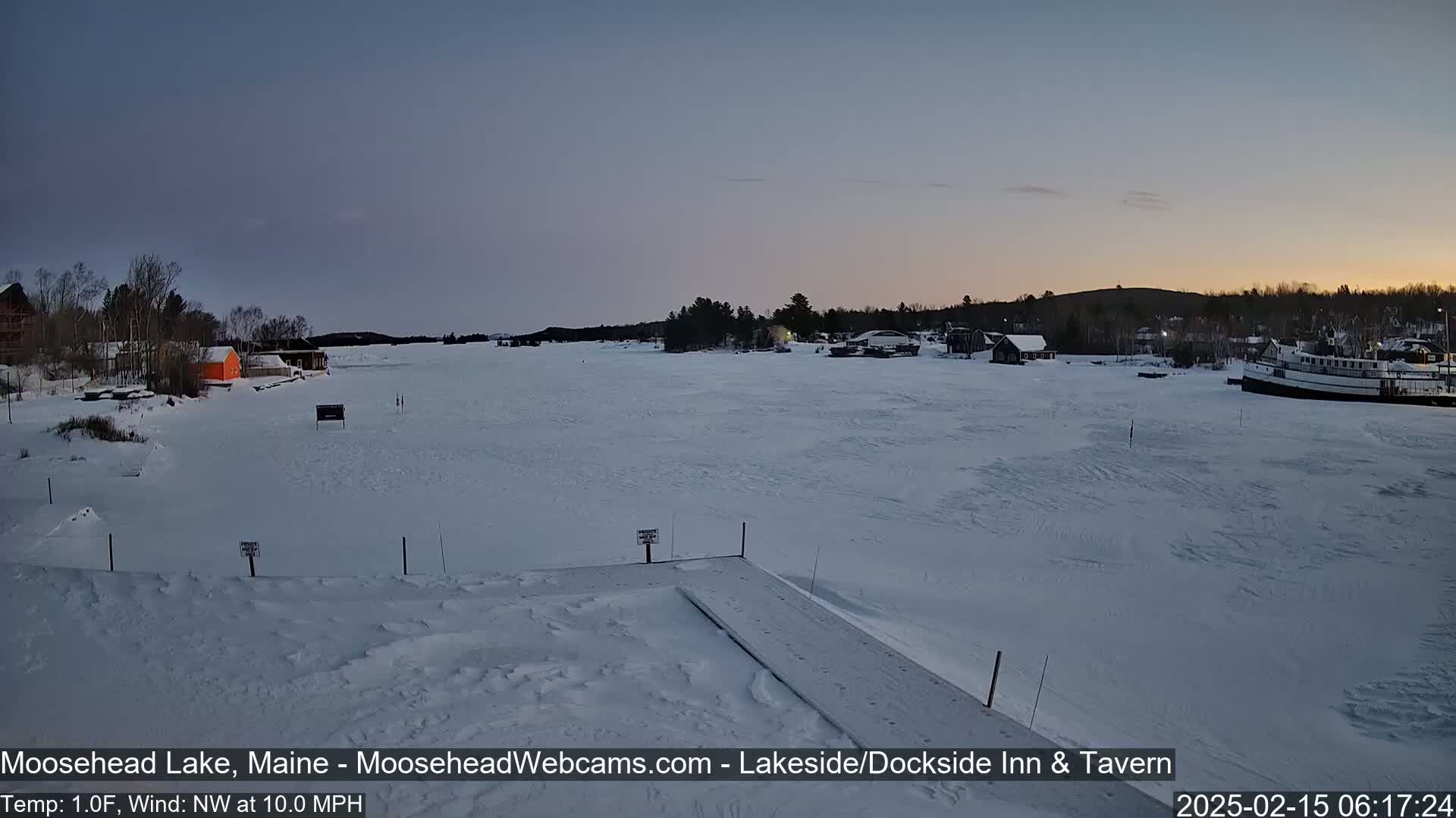 A frozen lake at dawn, with a few buildings and a boat visible on the shore, under a twilight sky and a 10 mph northwest wind.