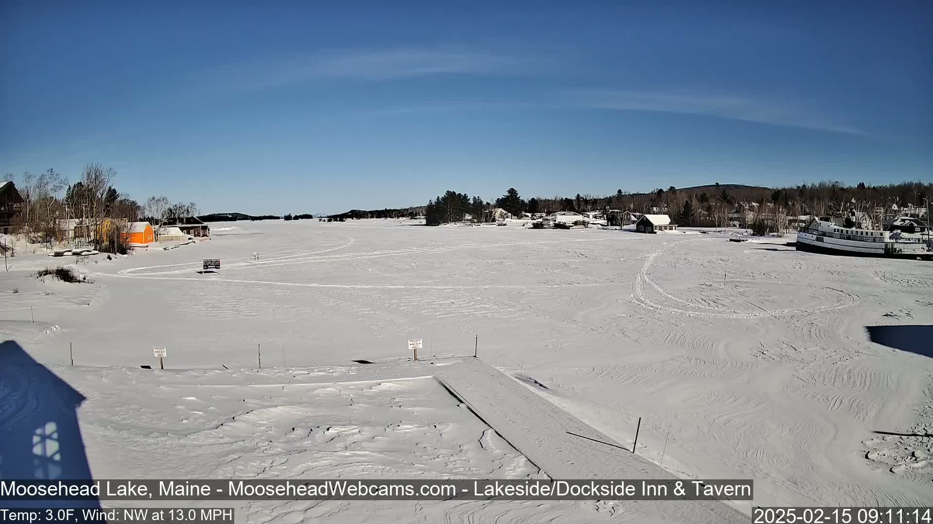 A snow-covered frozen lake is visible, with a few buildings and a boat on the shoreline under a clear blue sky.