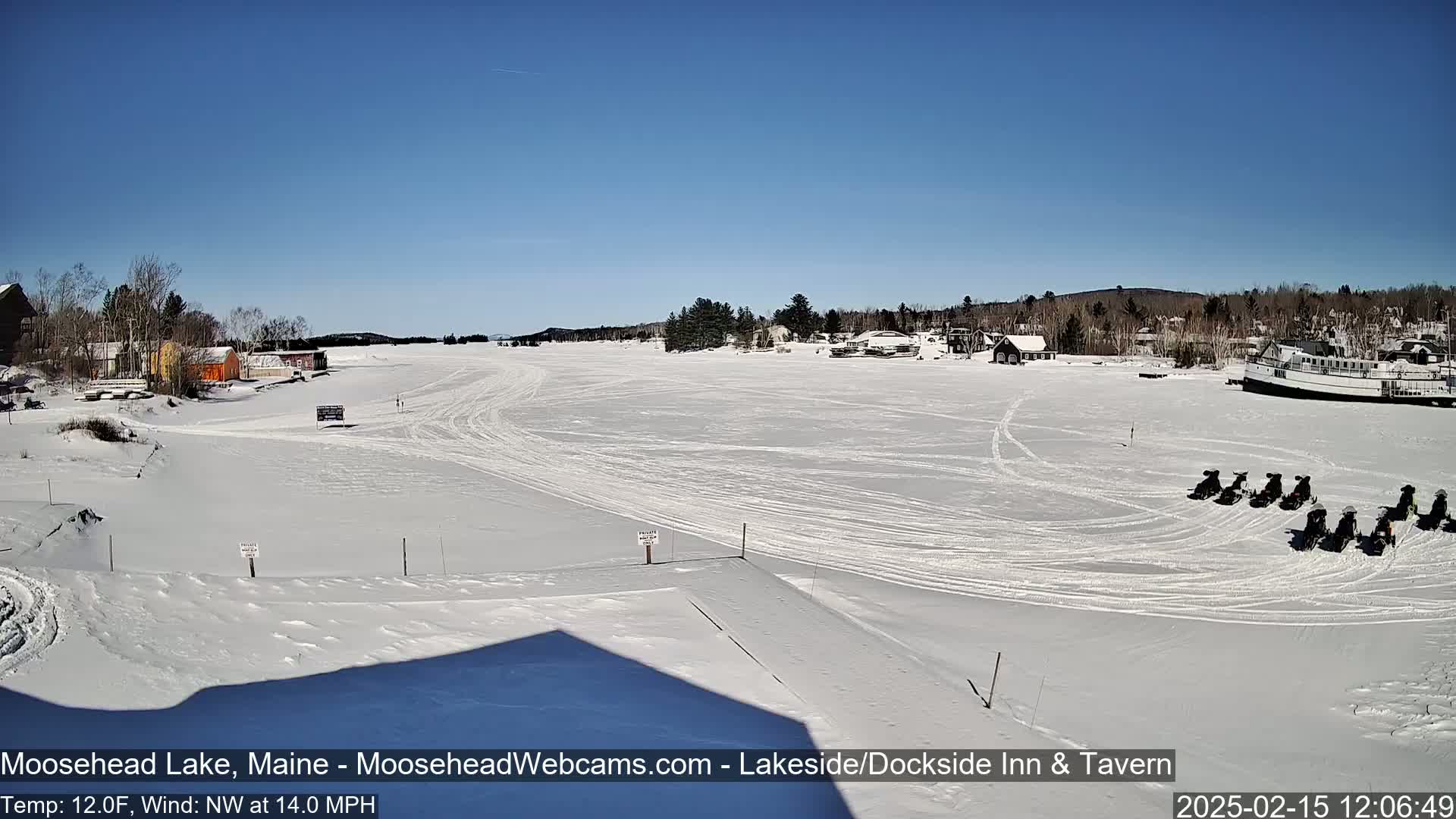 A snow-covered frozen lake with several snowmobiles parked on the ice, under a clear blue sky, and buildings along the shoreline.