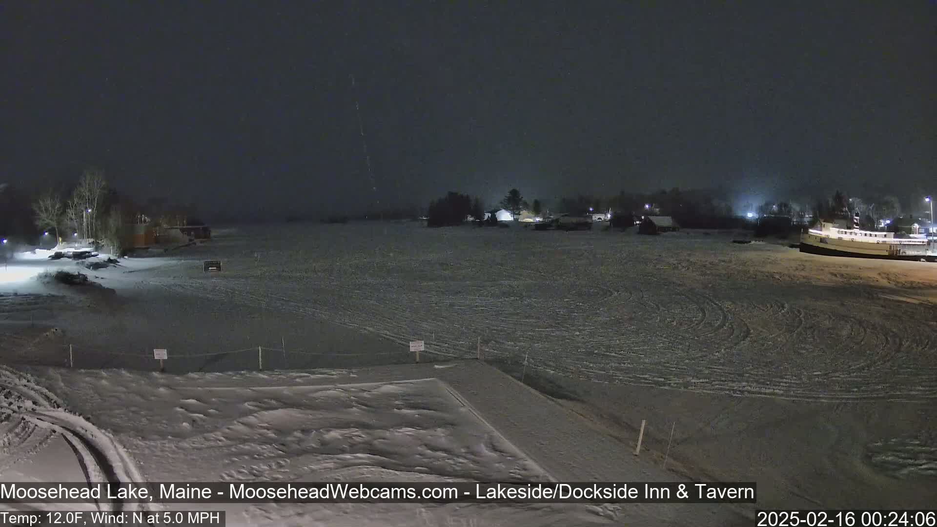 A snow-covered frozen lake at night, with a few buildings visible in the distance under a dark sky.