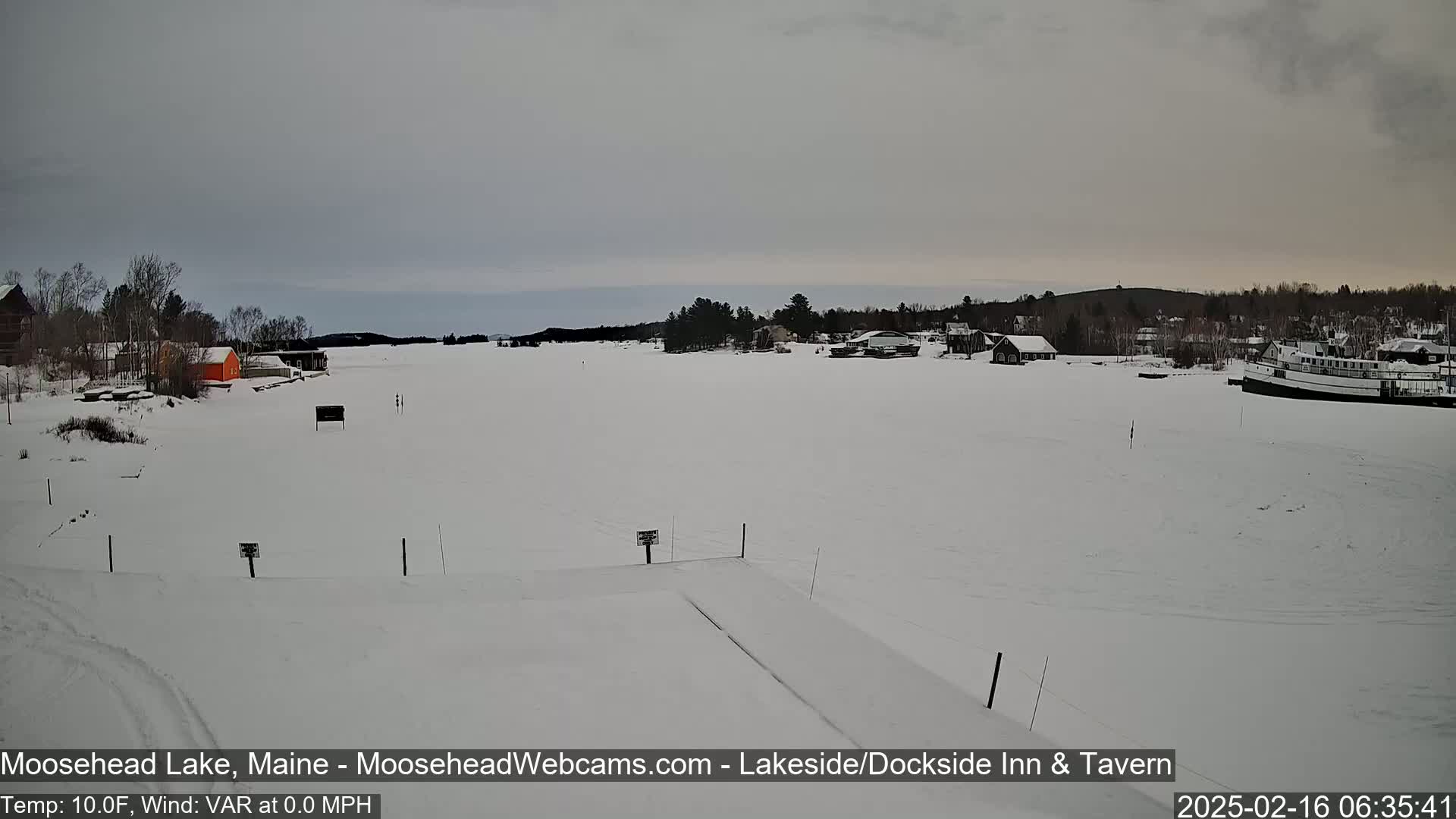 A snow-covered lake with buildings and a boat visible in the distance under an overcast sky.