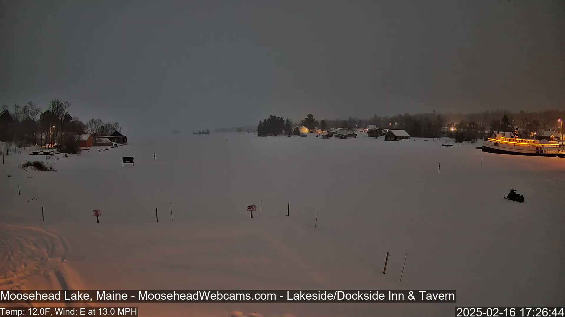 A snow-covered lake at dusk with a few buildings and a boat visible, under a dark, overcast sky with a wind from the east at 13 mph.