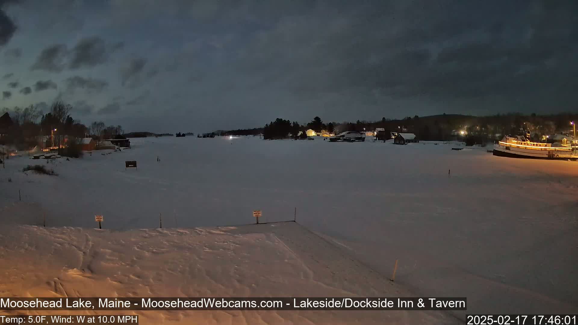 A snow-covered lake at night, with a few illuminated buildings and a large boat visible in the distance under a dark, cloudy sky.
