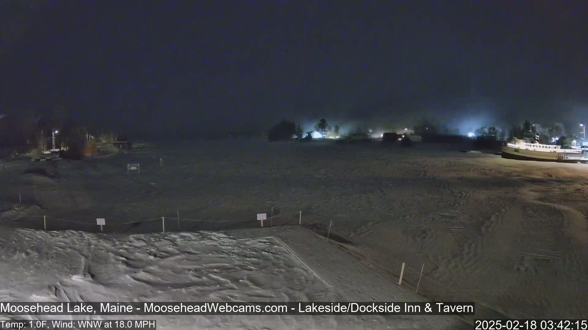 A snow-covered lakeside area is seen at night under foggy conditions, with buildings and lights faintly visible in the distance.