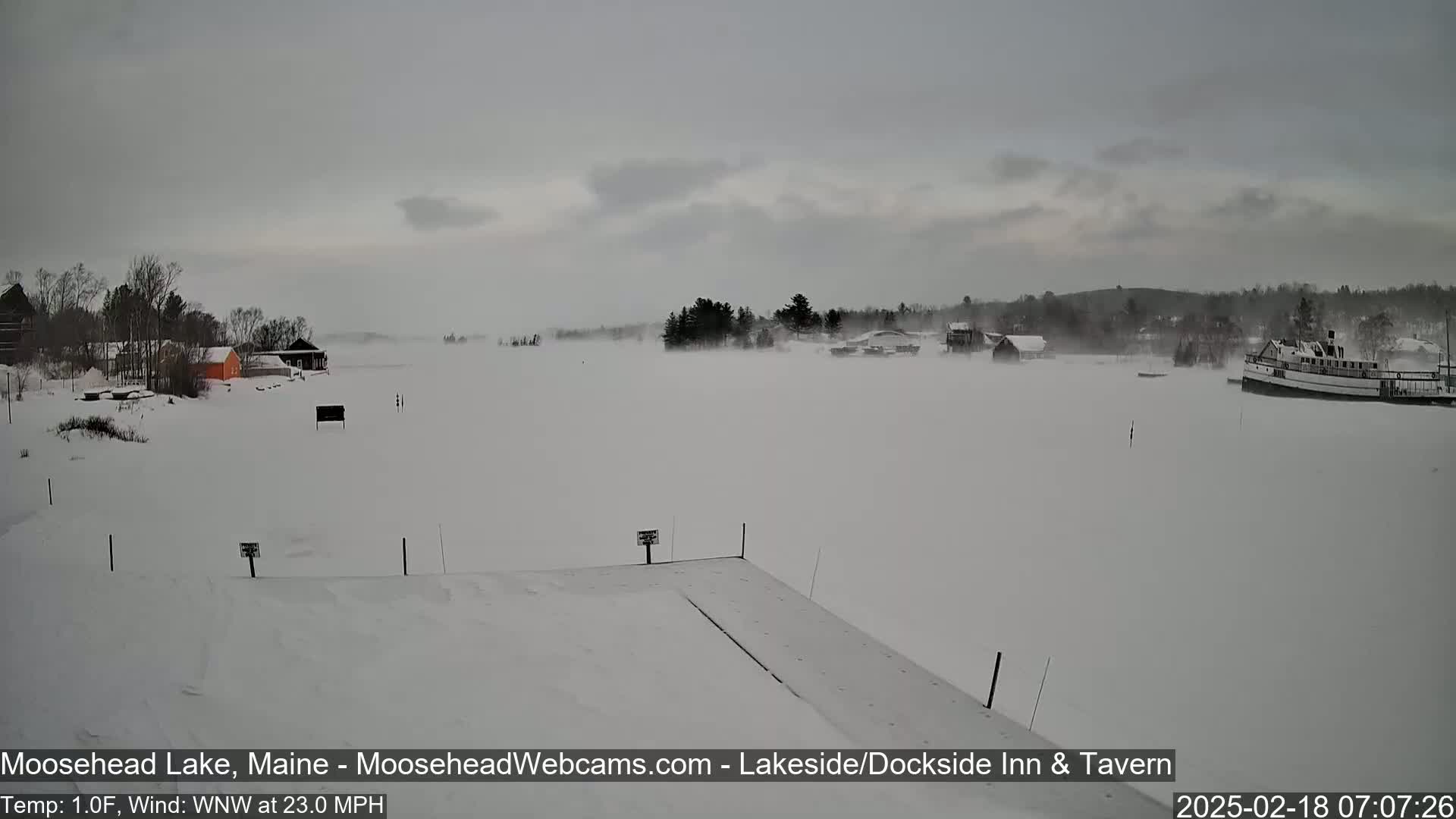 A snow-covered lake with a few buildings and a boat visible in the distance, under a cloudy sky with a strong wind.