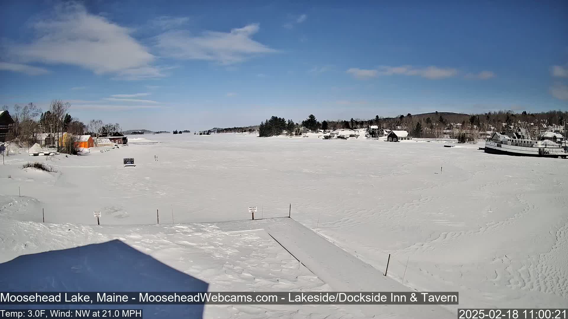 A snow-covered lake with buildings and a boat visible on the horizon under a mostly clear, sunny sky.