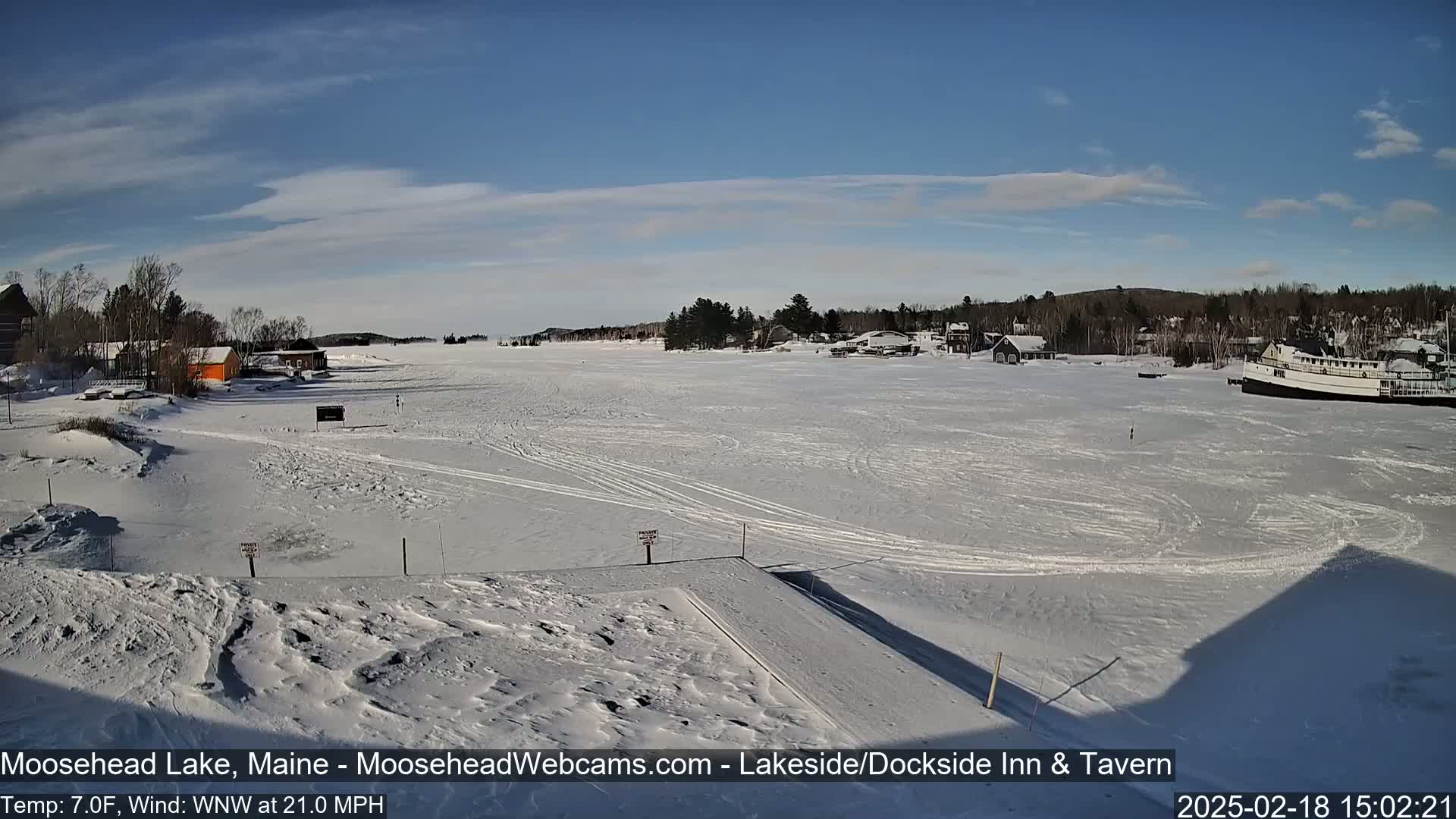 A frozen lake, under a mostly clear blue sky, shows a few buildings and a boat in the distance, with snow covering the ground and some tracks visible on the ice.