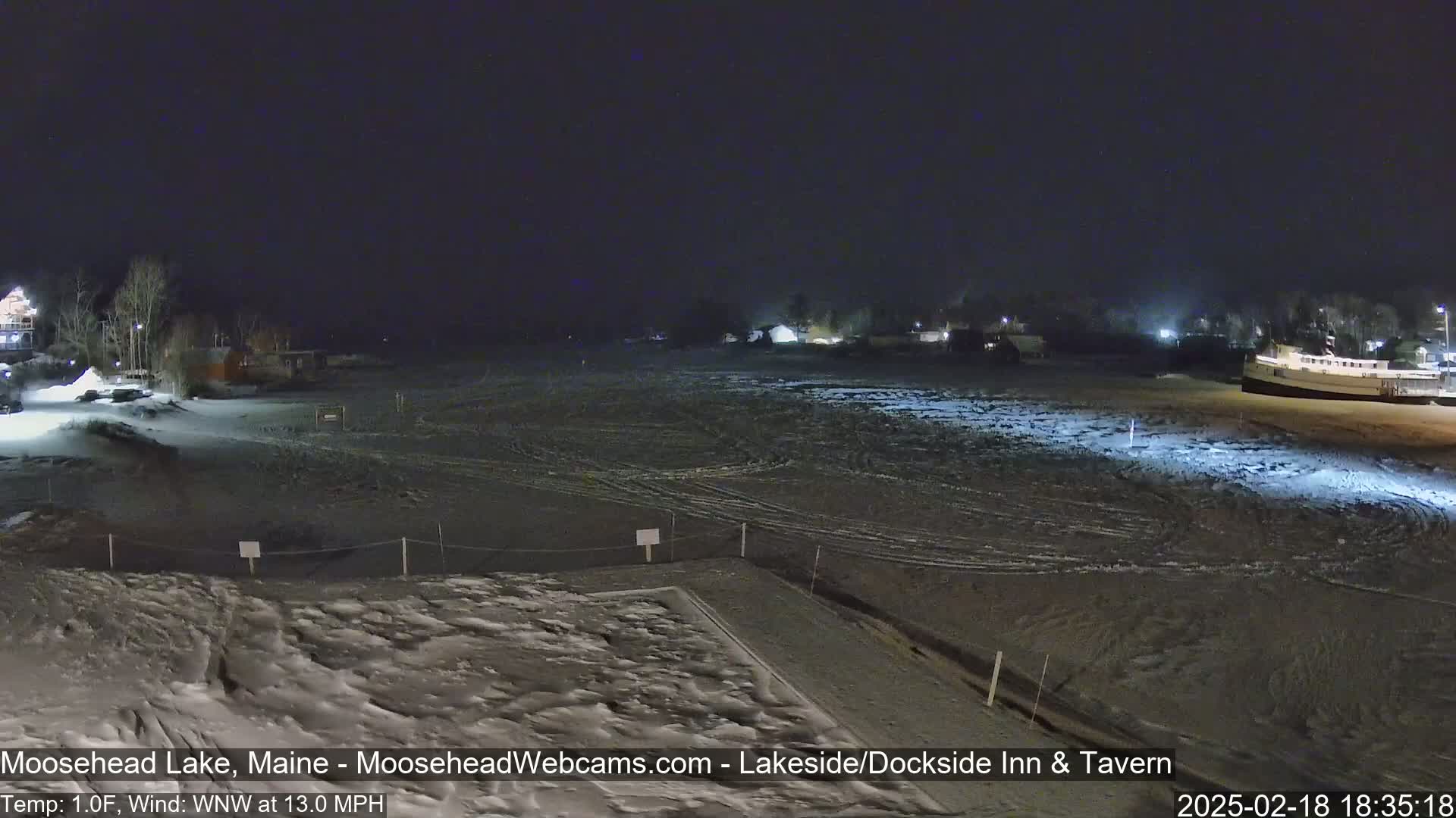 A snow-covered, dark nighttime scene shows a frozen lake with a few buildings and trees in the distance under a clear sky, with a wind of 13 mph.