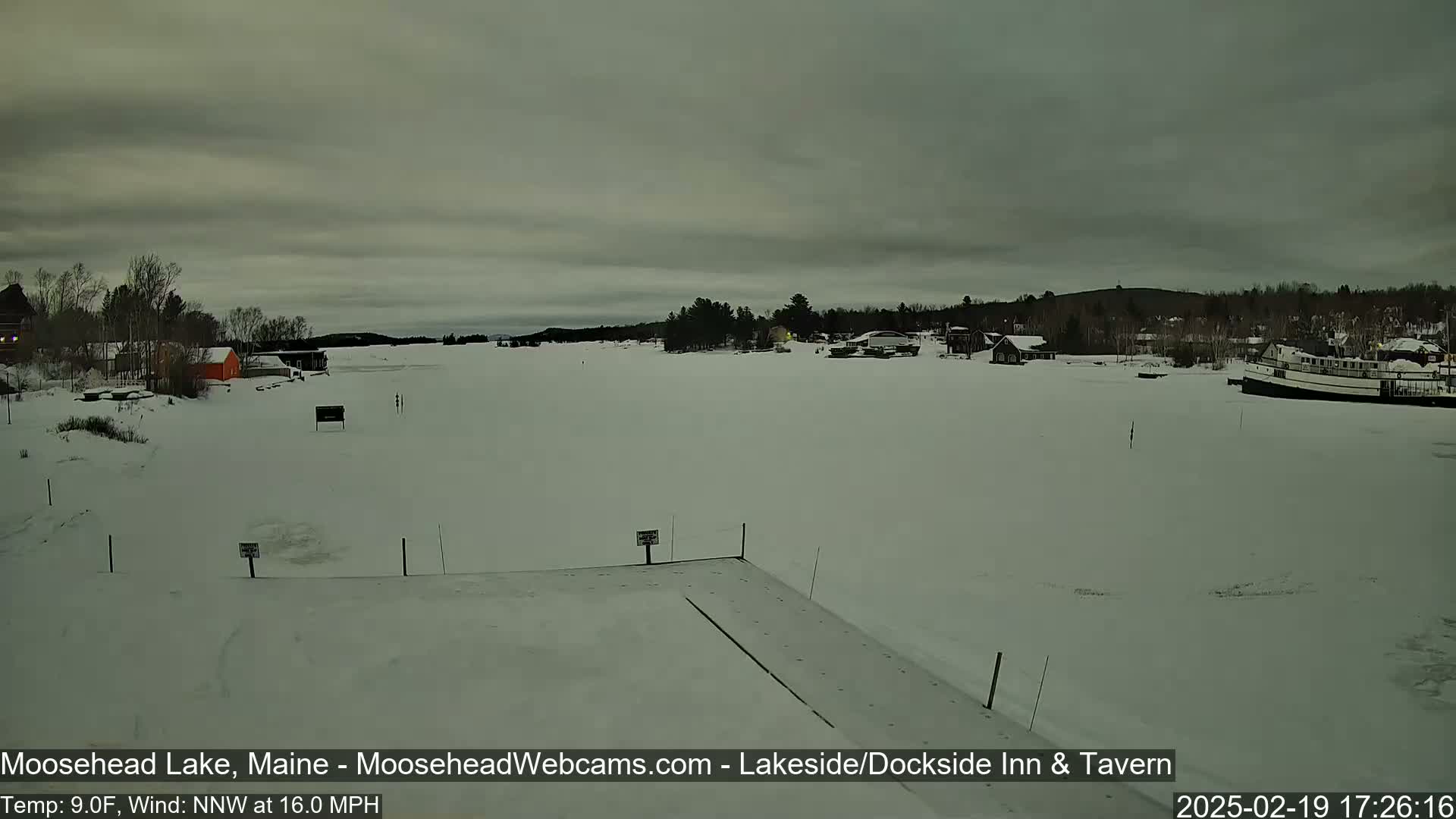 A frozen lake, under an overcast sky, shows a small town in the distance with a boat docked on the ice.