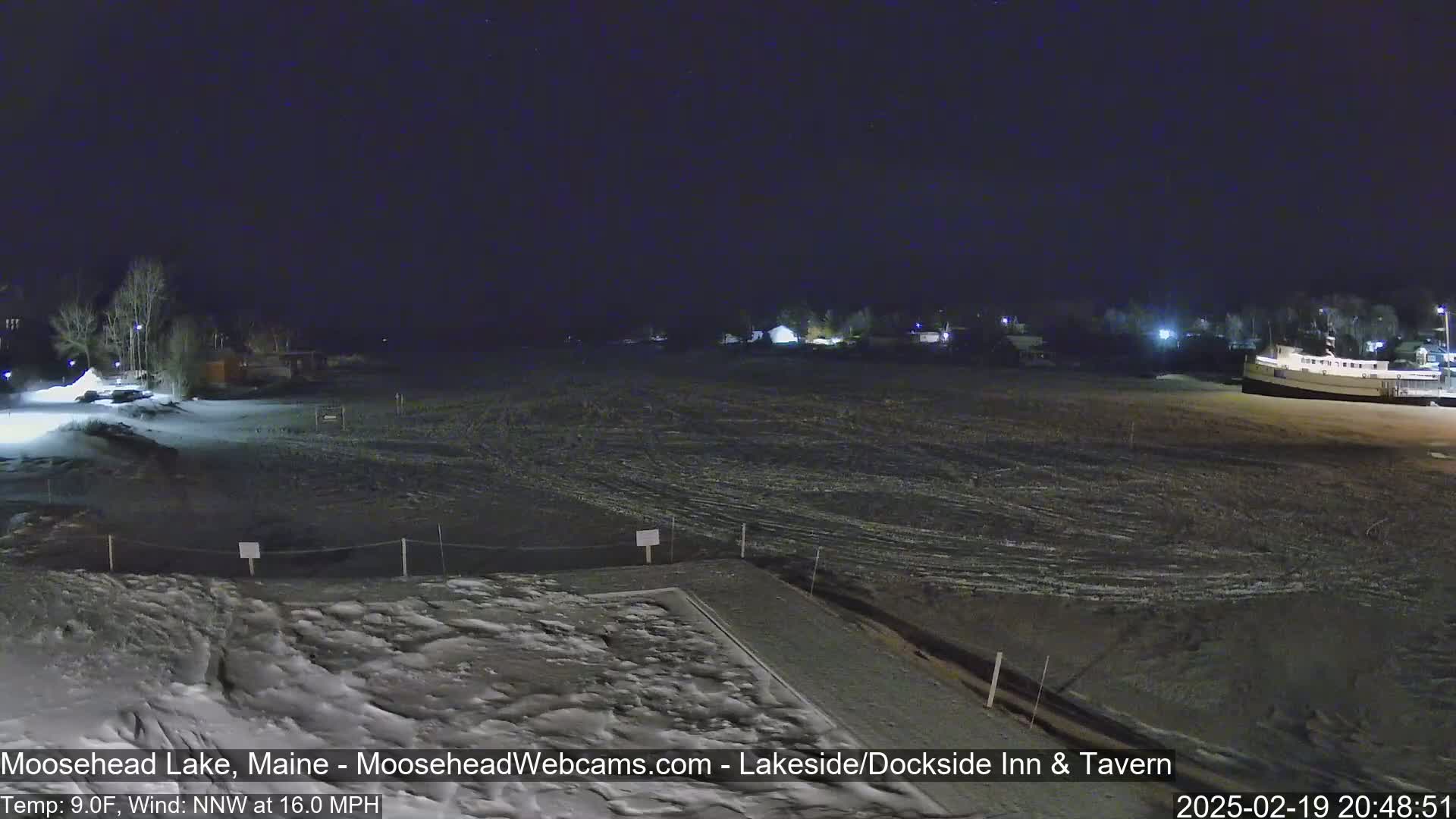 A snow-covered, nighttime lakeside scene shows a few buildings and a boat under a clear, dark sky with a north-northwest wind at 16 mph and a temperature of 9°F.