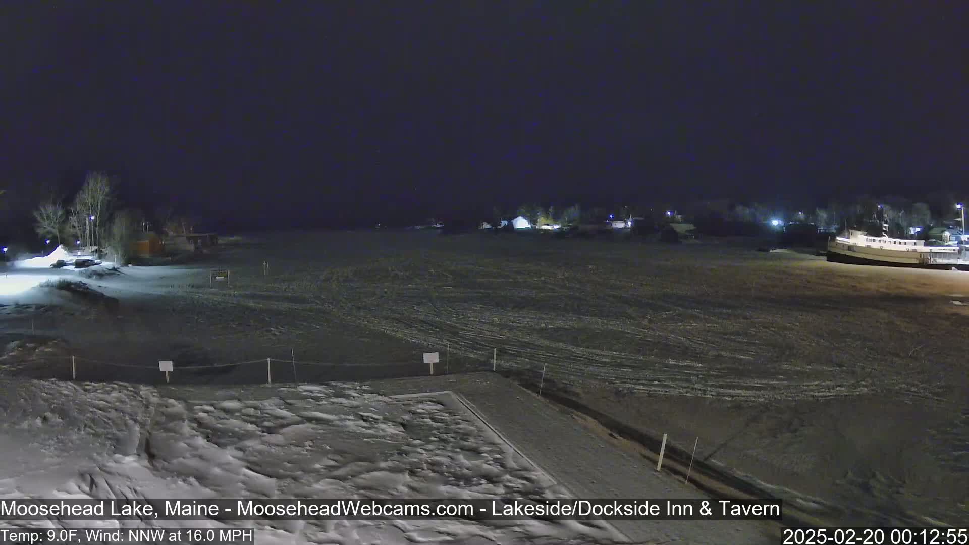 A nighttime view of a snow-covered lake shore with buildings and a boat in the distance under a dark sky, with a northwest wind at 16 mph and a temperature of 9°F.