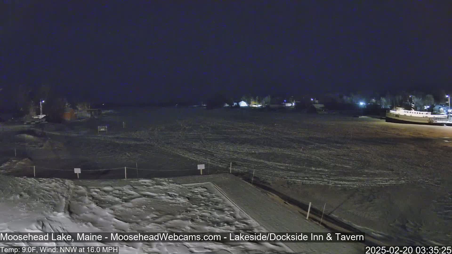 A nighttime view of a snow-covered lakefront area with a building and a boat visible in the distance under a dark sky, with a northwest wind blowing at 16 mph and a temperature of 9°F.