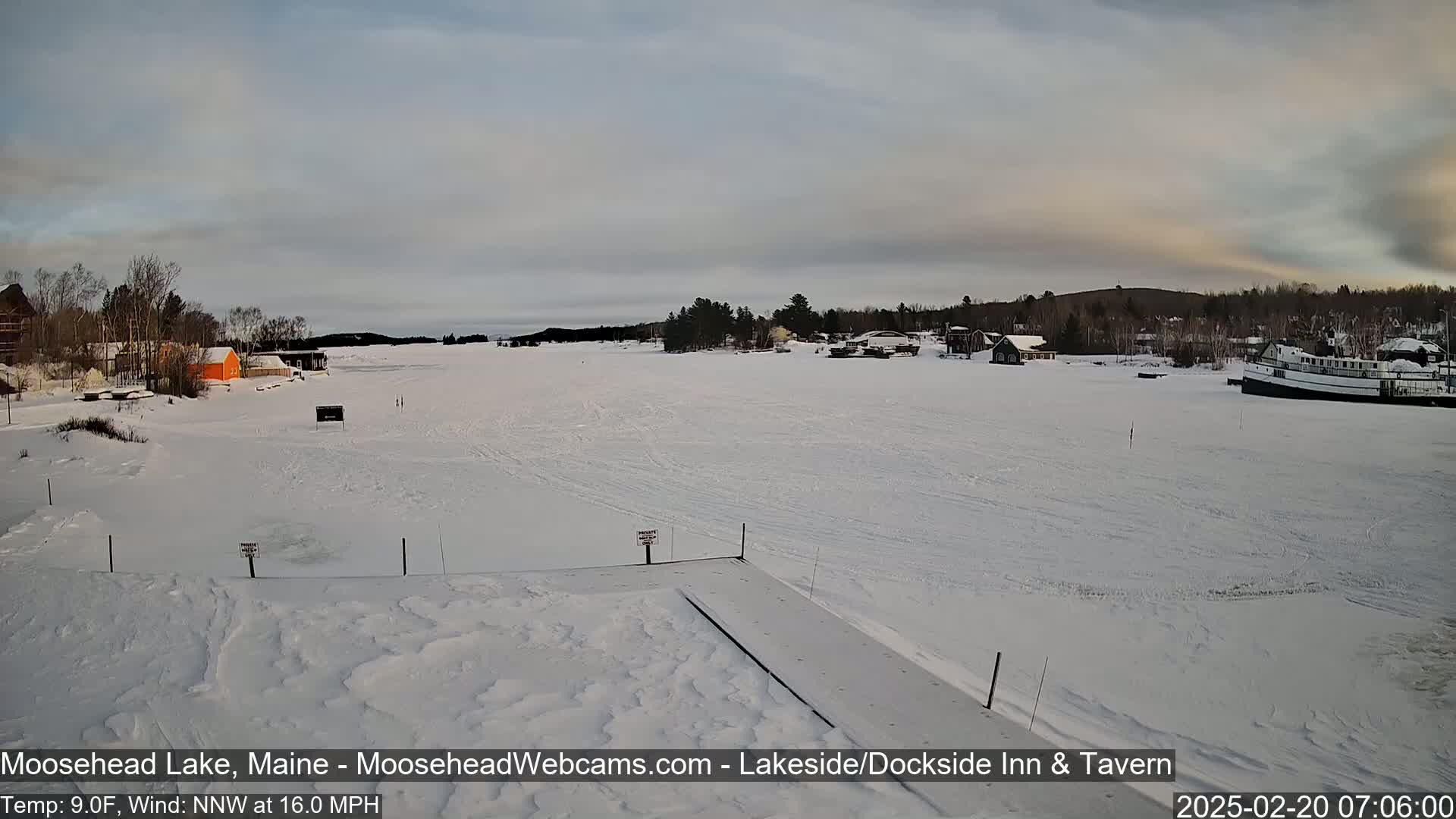 A snow-covered frozen lake with a few buildings and a boat visible in the distance, under a mostly cloudy sky.