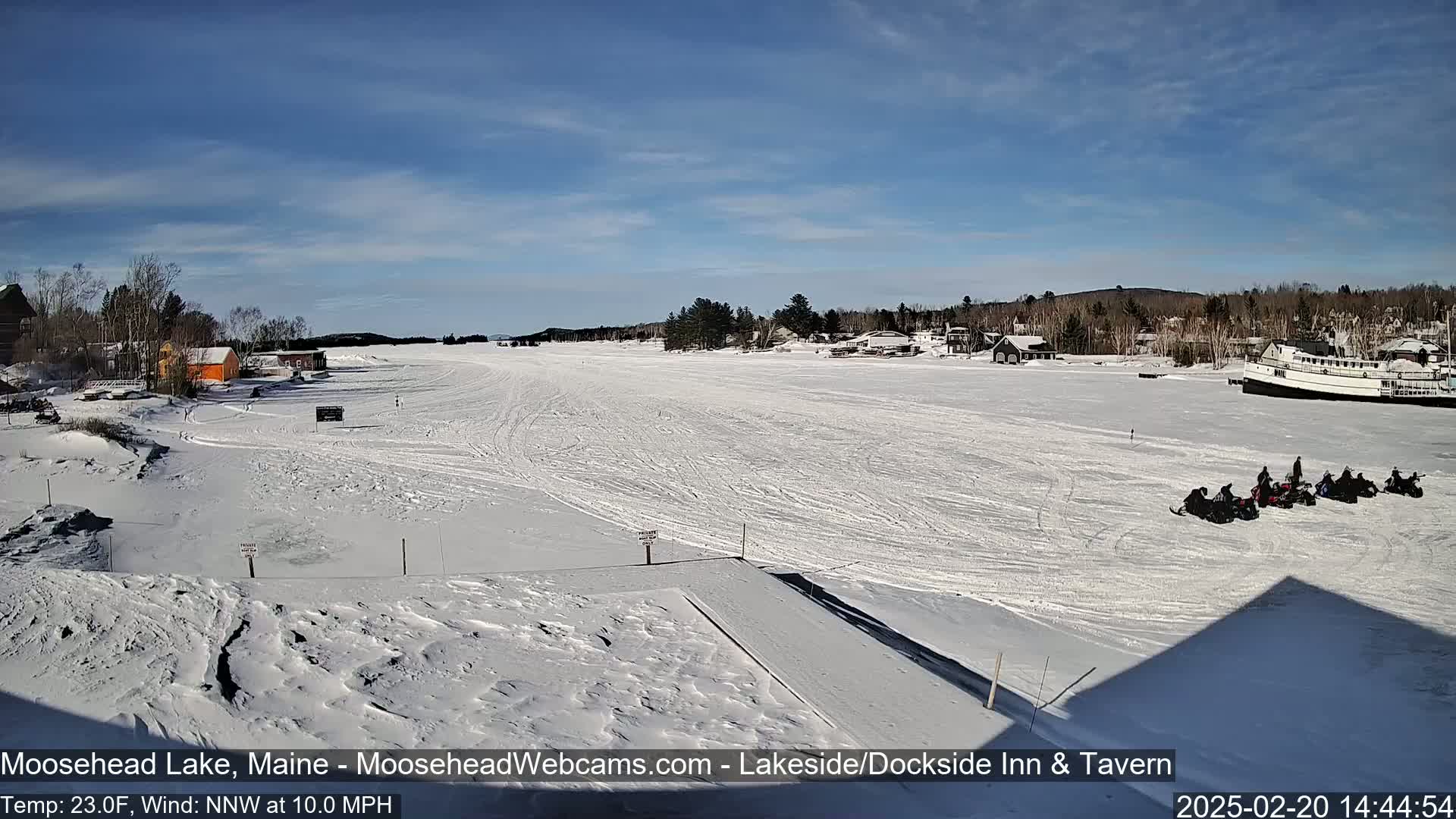 A snow-covered lake, under a mostly sunny sky, features several snowmobiles parked near a shoreline with buildings and a boat.