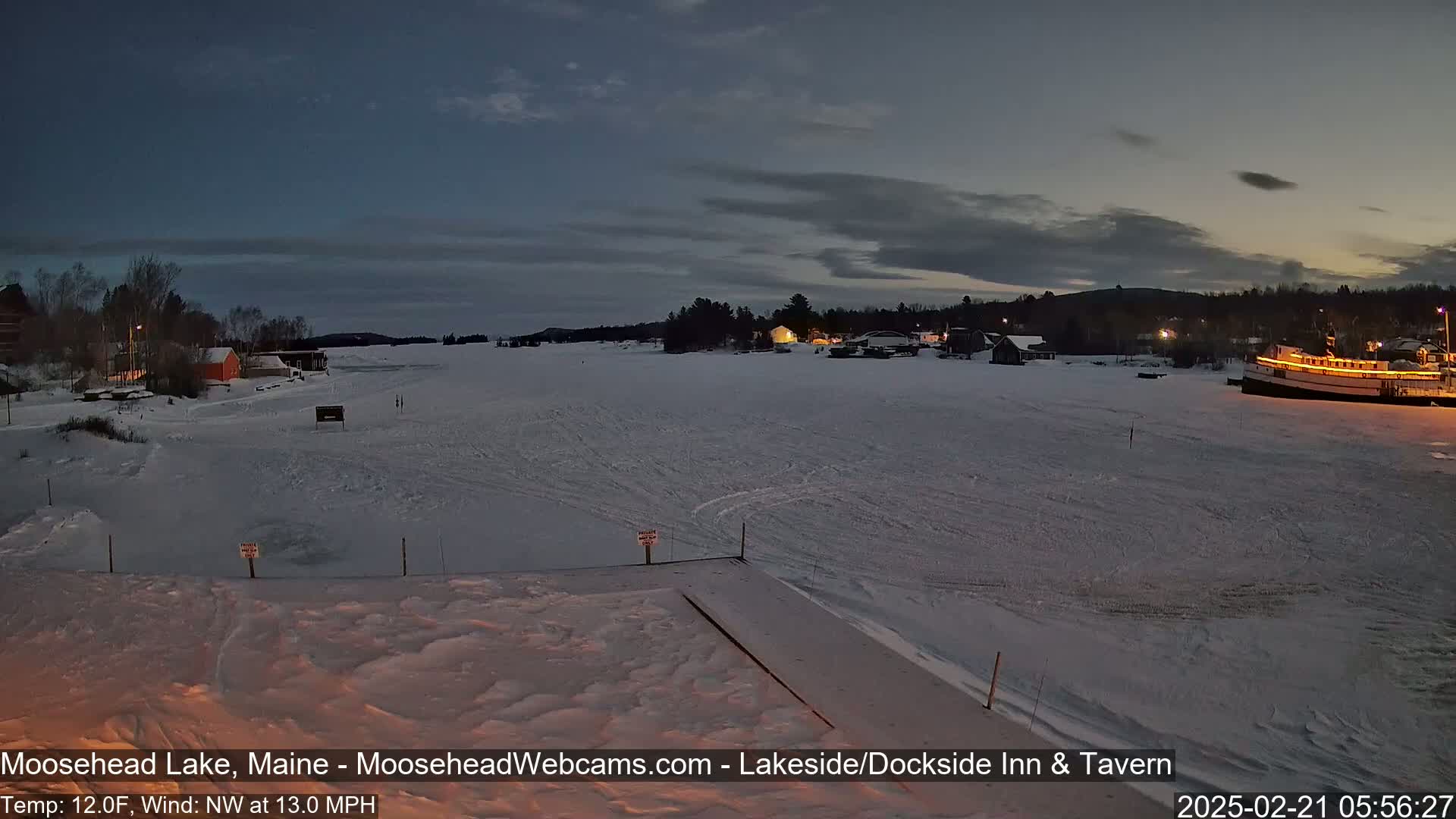A frozen lake at twilight, with a few buildings and a boat visible on the shore under a mostly clear sky.