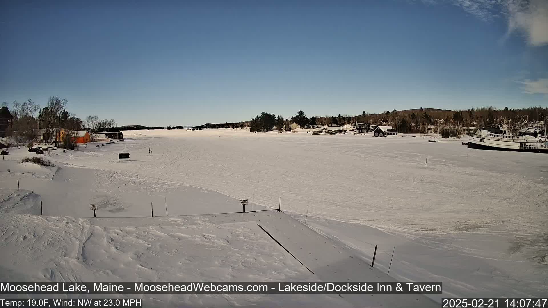A frozen lake, under a clear blue sky, features a few buildings along the shore and a boat docked on the ice.