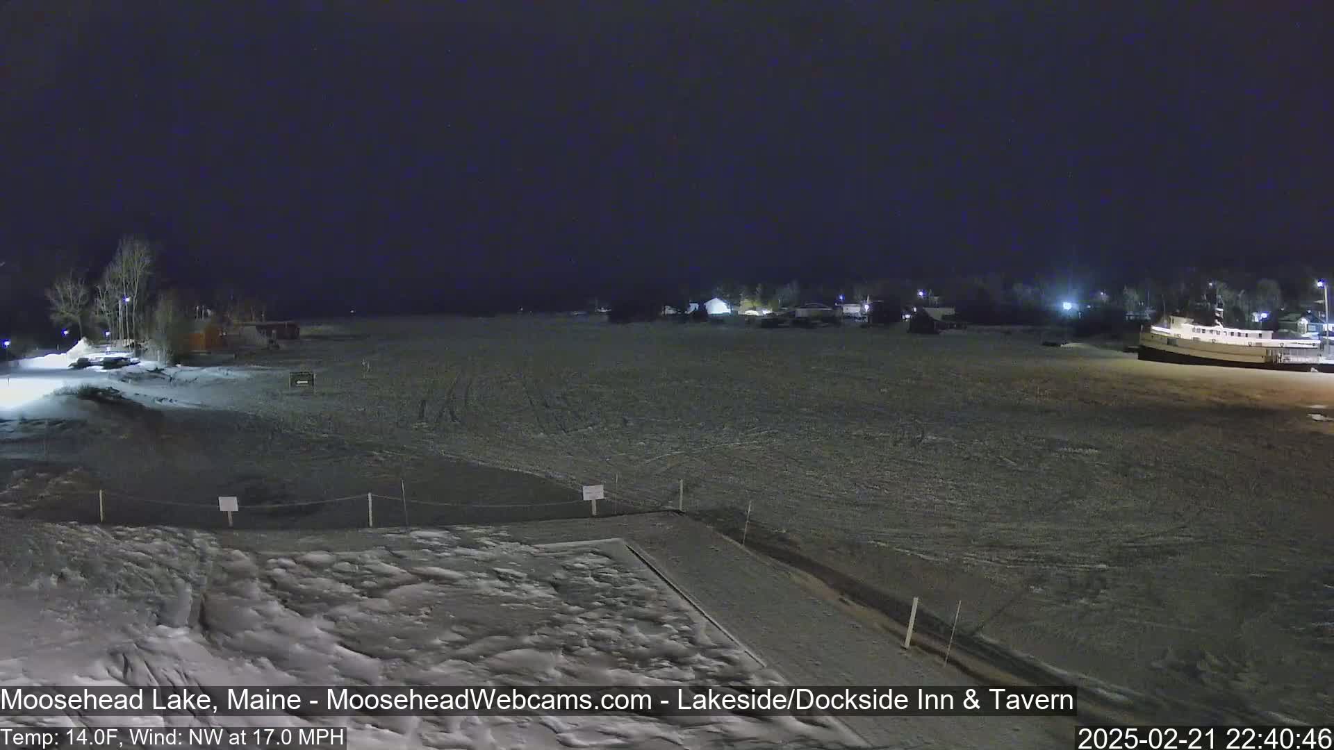 A snow-covered lakeside area at night with a few buildings and a boat visible under a dark sky, with a northwest wind at 17 mph and a temperature of 14 degrees Fahrenheit.