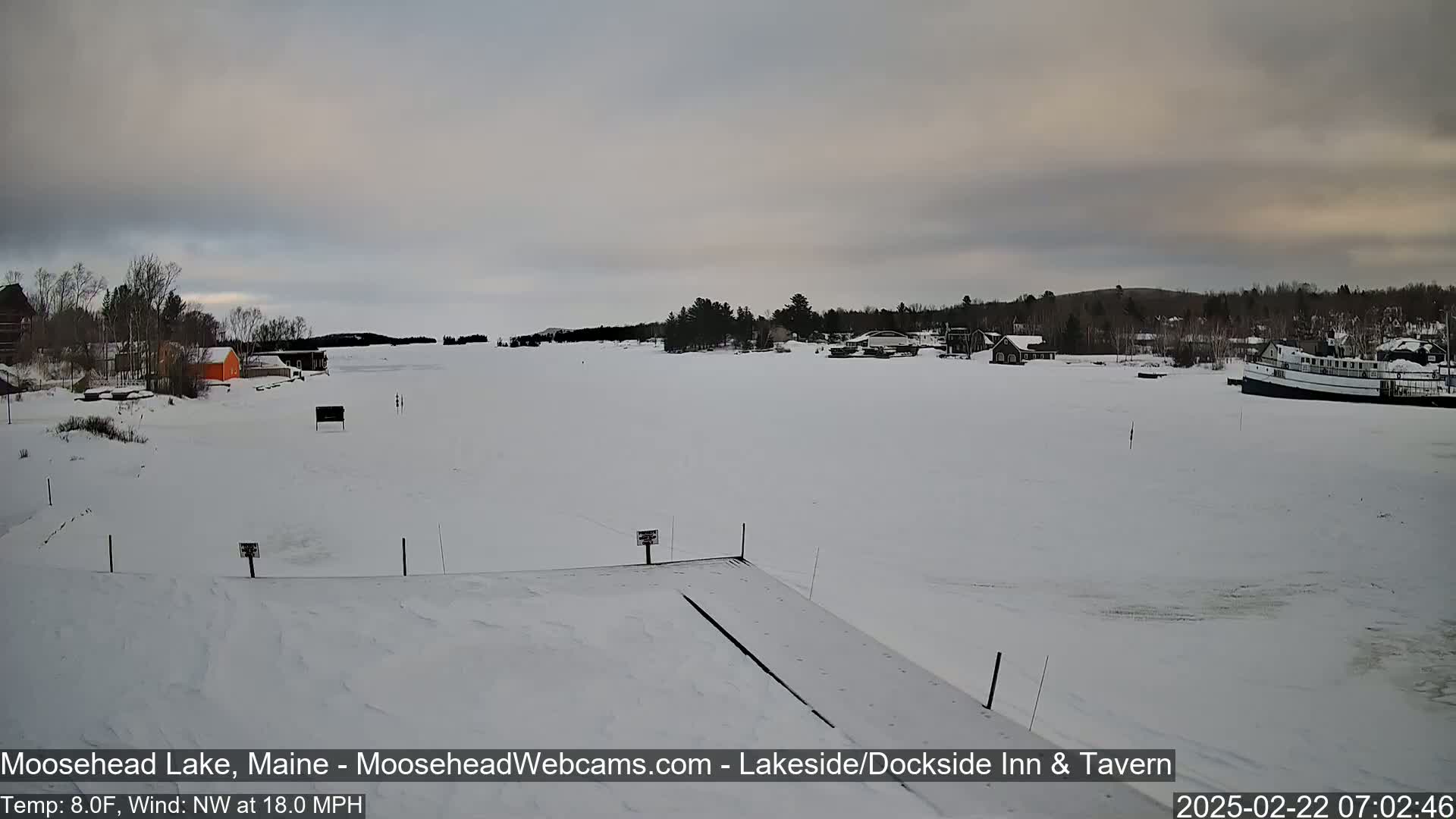 A snow-covered frozen lake with buildings and a boat visible in the distance under a cloudy sky.