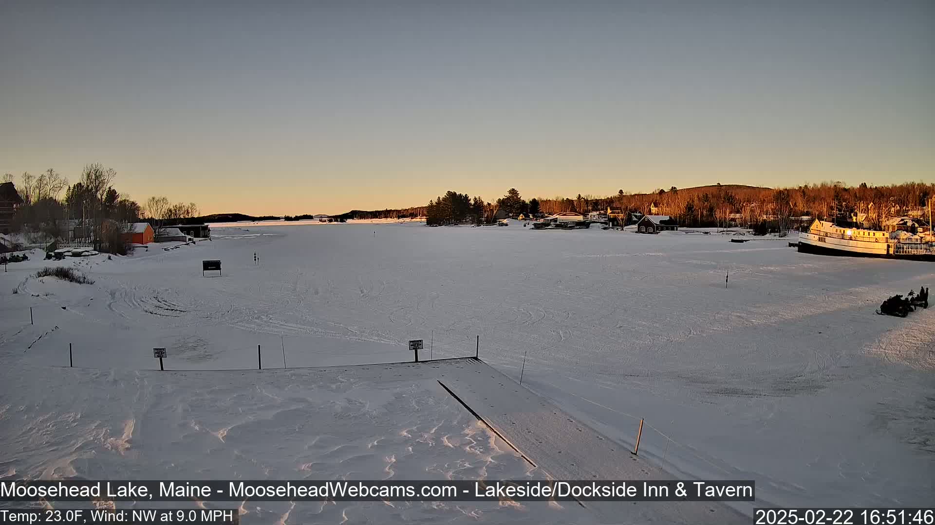 A frozen lake at sunset, with a few buildings and a boat visible on the shore, under a clear sky and a northwest wind of 9 mph at 23 degrees Fahrenheit.