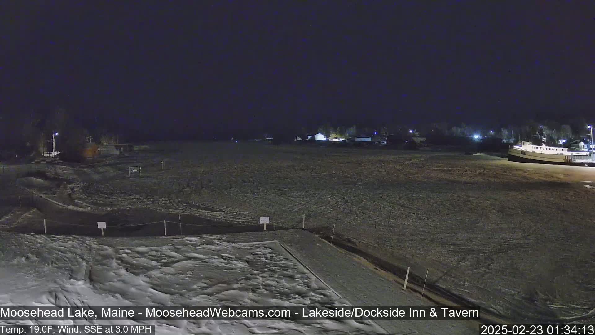 A nighttime view of a snow-covered lakeside area with a boat docked, under a clear, dark sky, and a light wind.