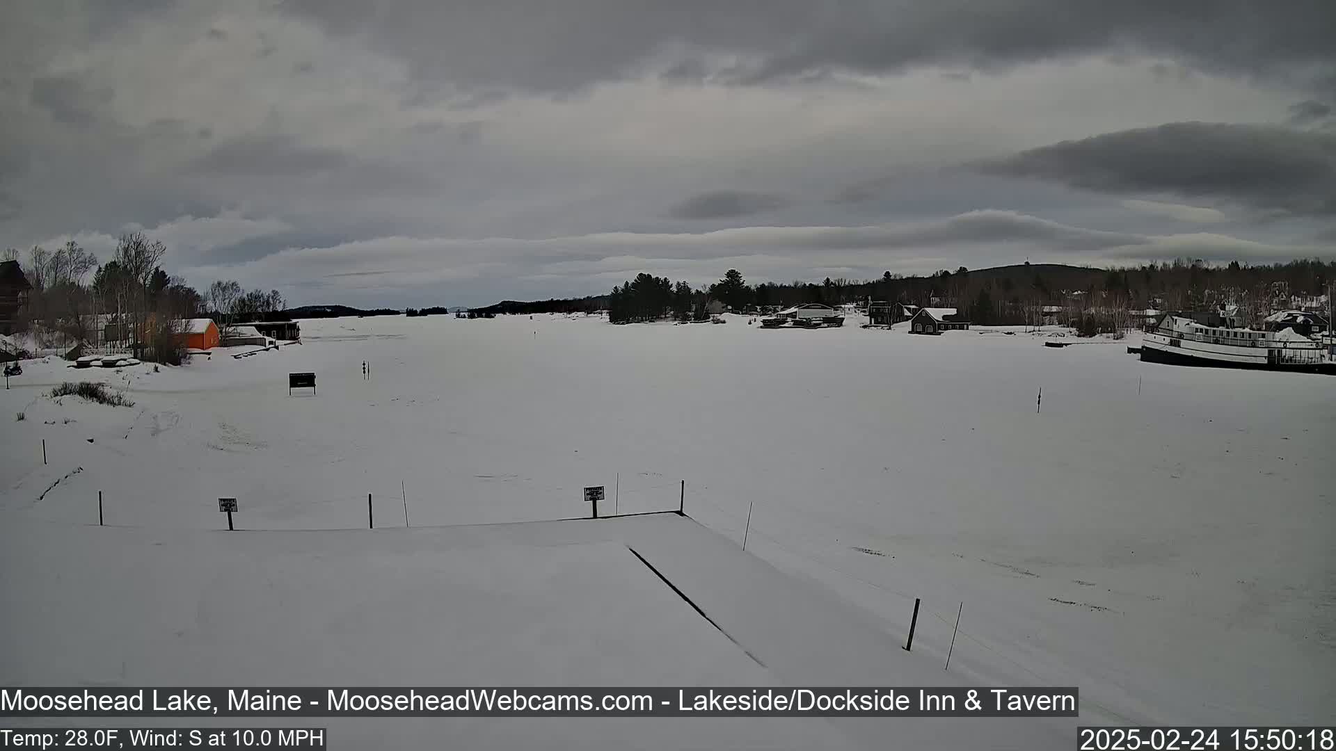 A snow-covered frozen lake is seen under an overcast sky, with a few buildings and a boat visible in the distance.