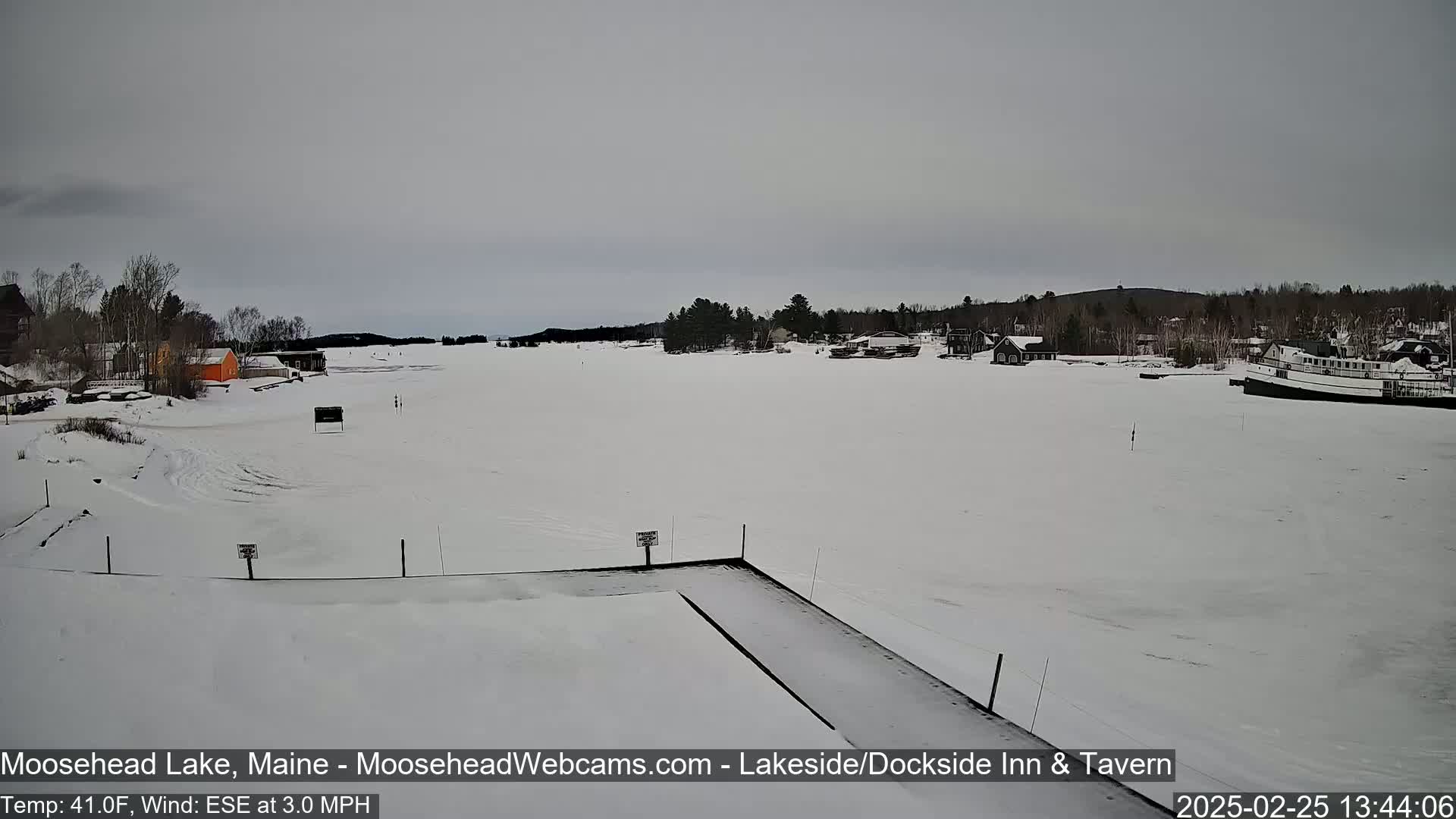 A snow-covered lake stretches out before a shoreline with buildings and trees under a cloudy sky.